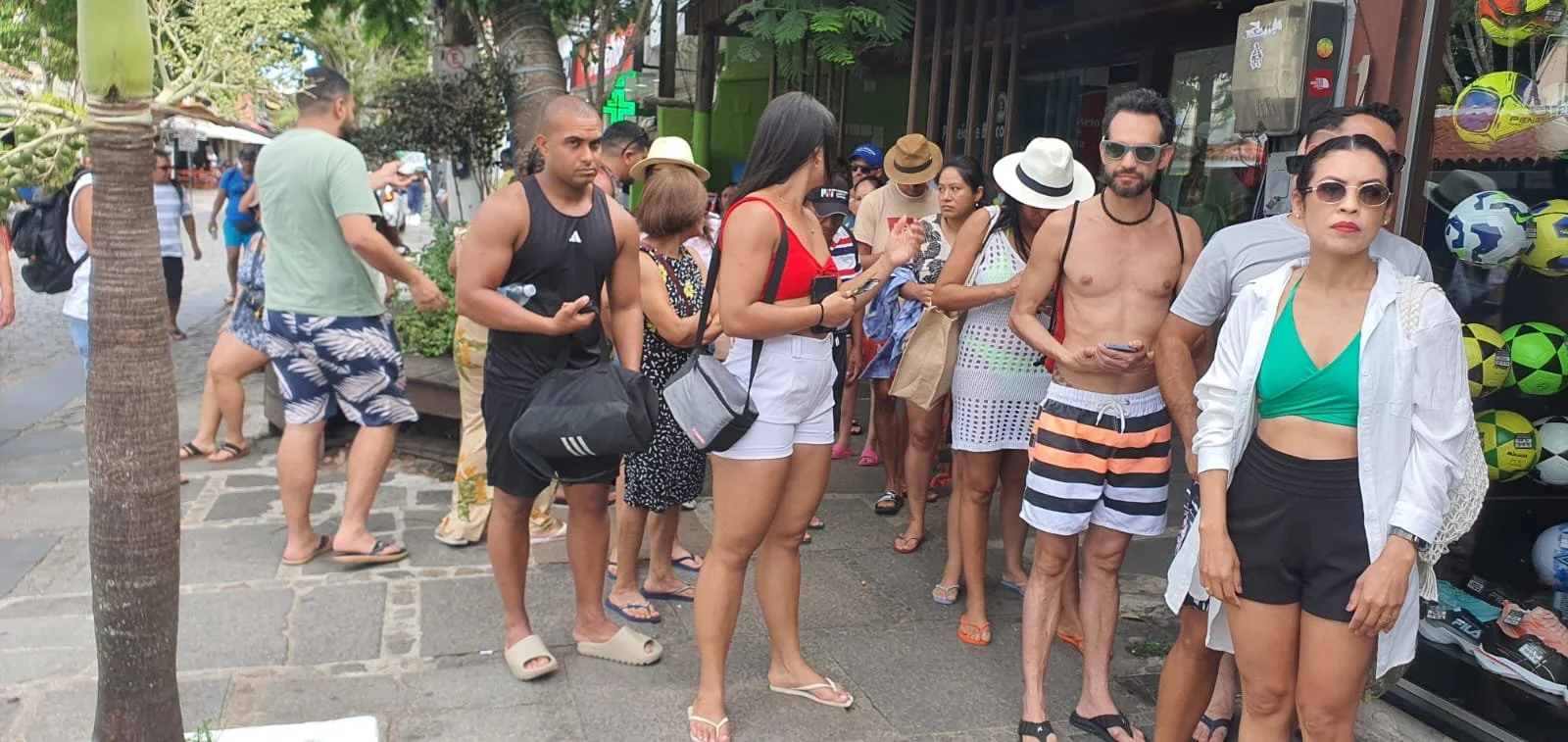 Pessoa com camiseta branca e cabelo longo escuro, com óculos escuros, posicionada na frente de uma loja de bolas de futebol, ao lado de uma pessoa com chapéu branco, camiseta cinza e shorts preto, ao fundo há várias pessoas na fila e árvores na rua.