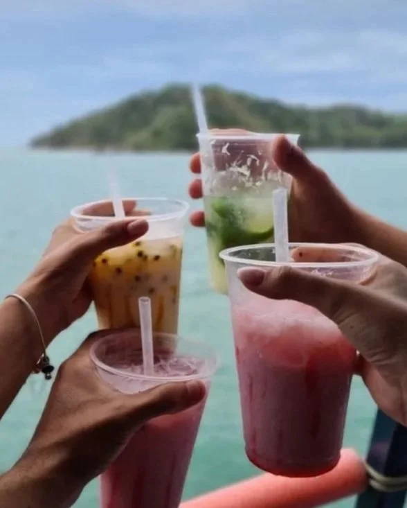 Quatro copos de smoothies de frutas sendo segurados por mãos, com um cenário de mar e ilha ao fundo.