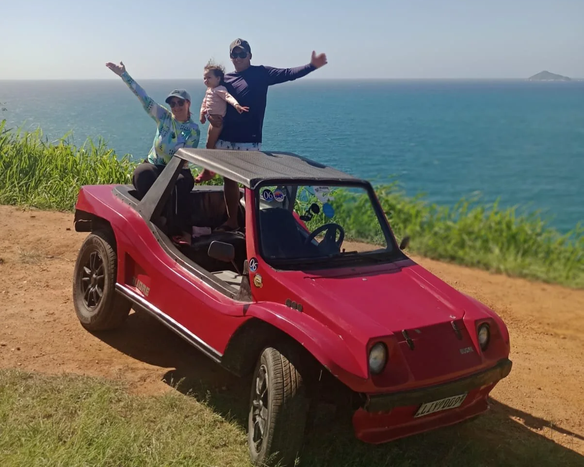 Três pessoas em um carro de brinquedo vermelho em um local à beira-mar com vista para o oceano, uma mulher, um homem e uma criança, comemorando ou posando para a foto.