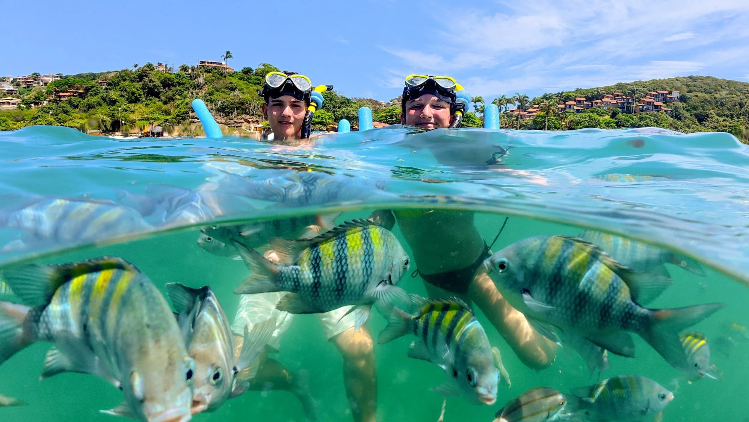 Duas crianças com equipamentos de mergulho na água, observando peixes coloridos, com uma área residencial no fundo e céu claro.