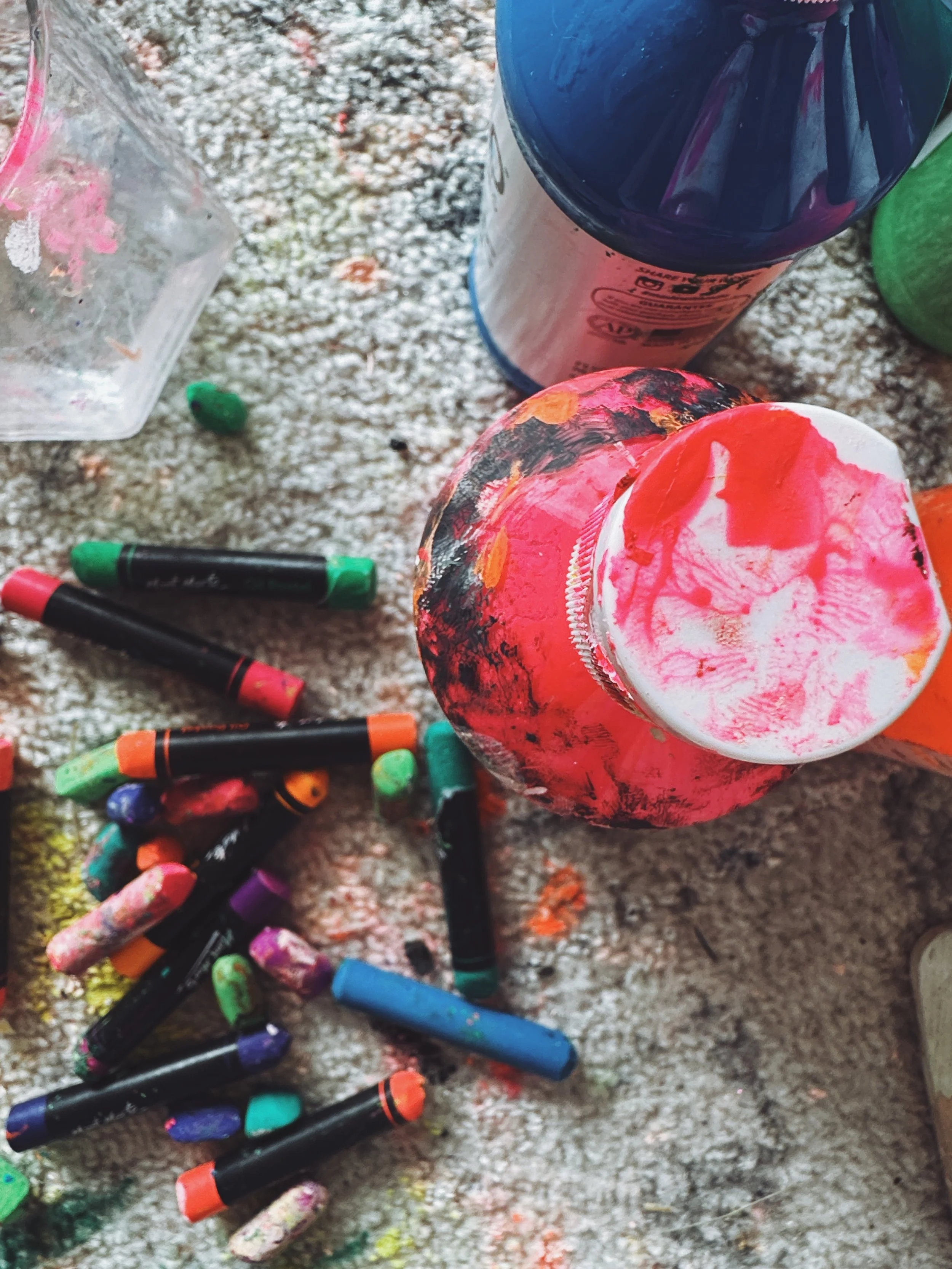 A collection of colorful crayons scattered on a dusty, textured surface with a partially opened container of pink and white paint nearby.