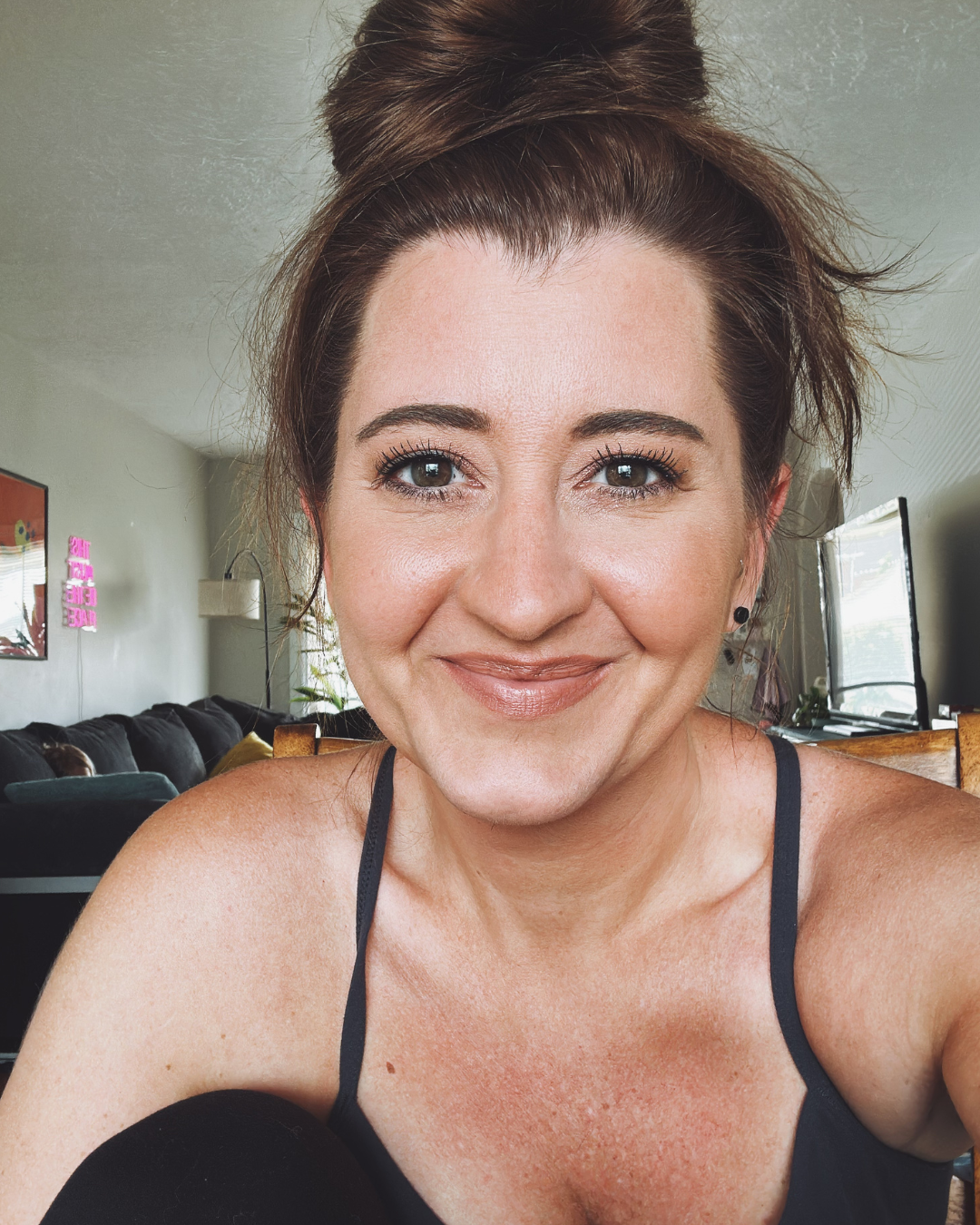Close-up of a smiling woman with brown hair in a bun, wearing black spaghetti straps top, in a living room with a couch, a wall decor, and a television in the background.