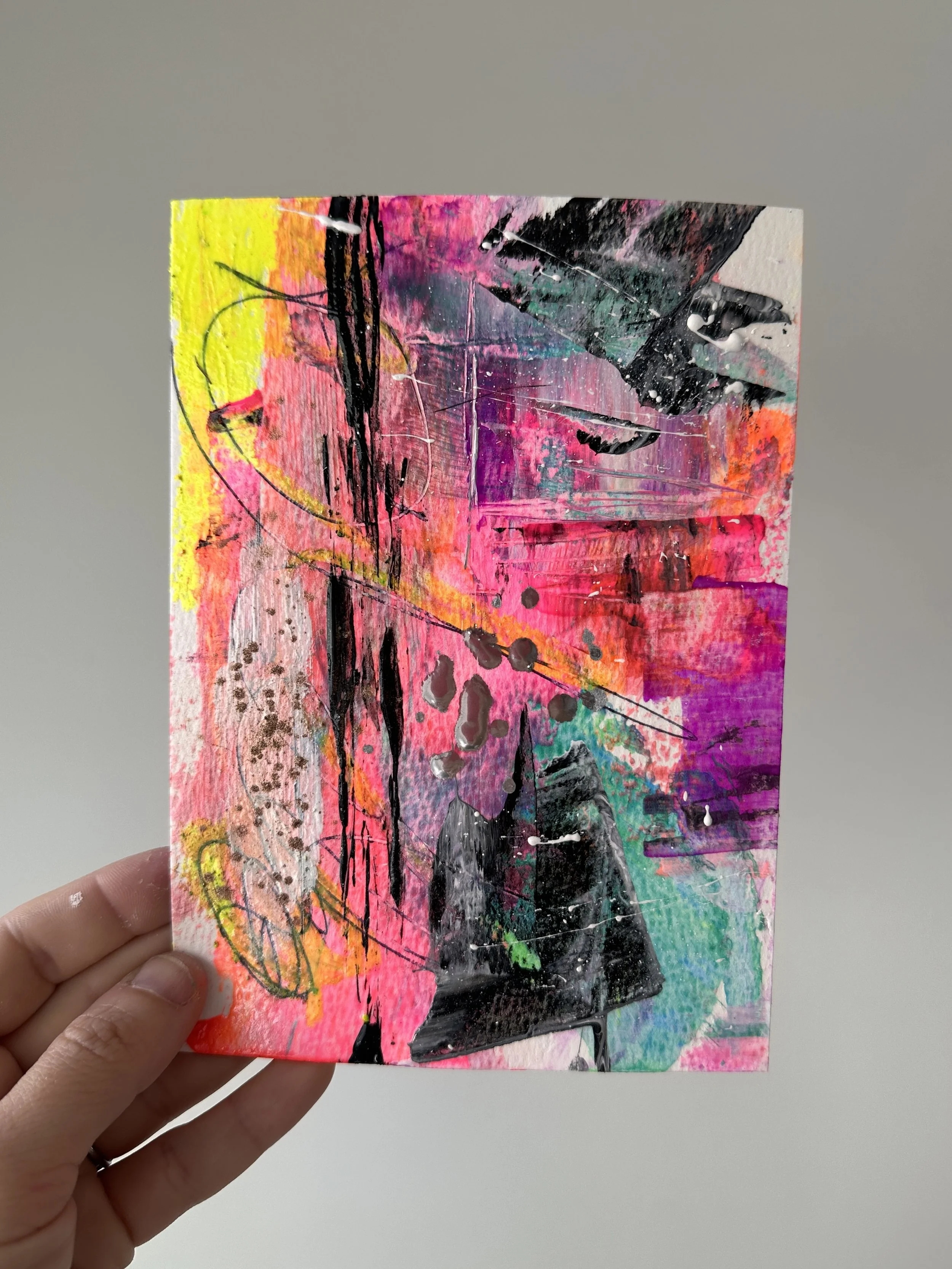 Abstract colorful painting with pink, yellow, black, purple, and green paint splatters and streaks, held by a person's hand.