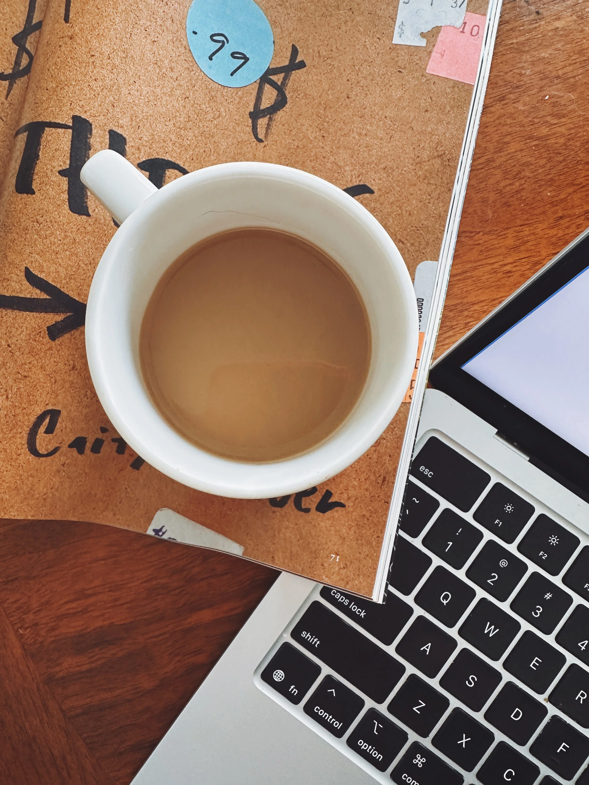 A white coffee mug filled with coffee placed on a wooden table. Part of a laptop with a black keyboard is visible in the bottom right corner. A brown paper notepad with handwritten notes and small sticky notes is underneath the mug.
