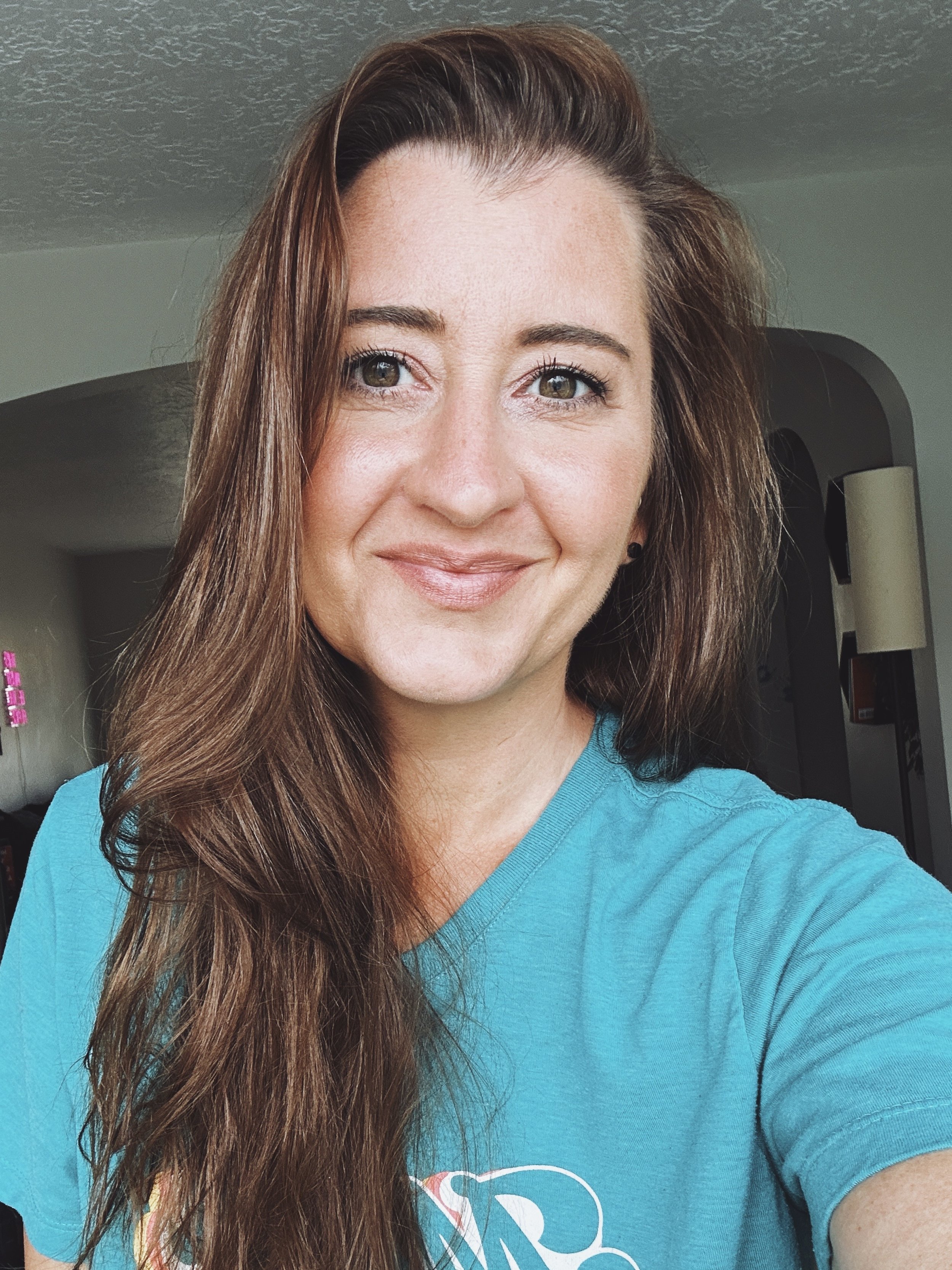 Close-up of a woman with long brown hair wearing a blue shirt, smiling, standing in a room with a curved doorway and a lamp in the background.