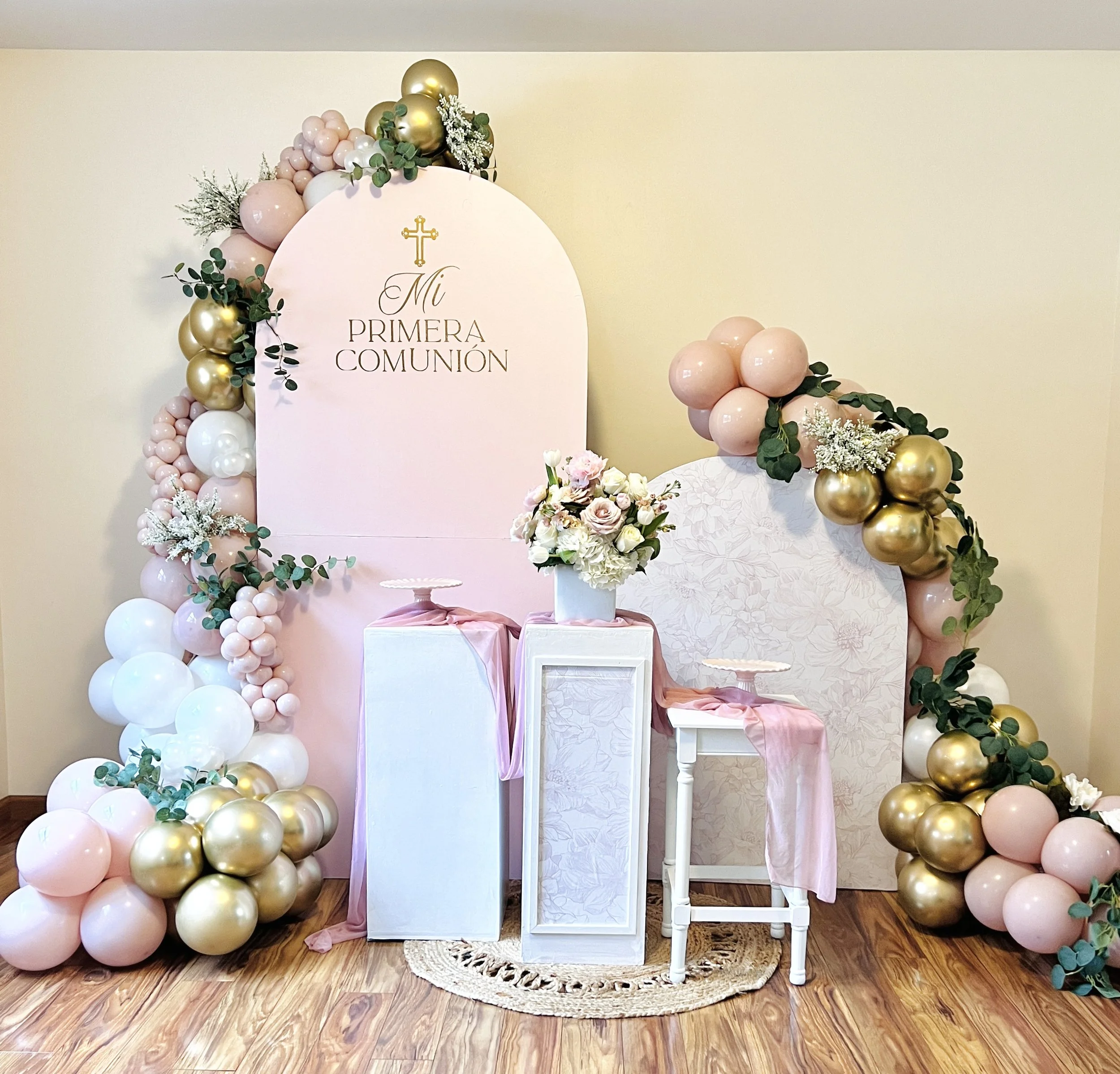Decorative backdrop for a First Communion celebration with pink, white, and gold balloons, floral arrangements, and a sign that reads "Mi Primera Comunión."
