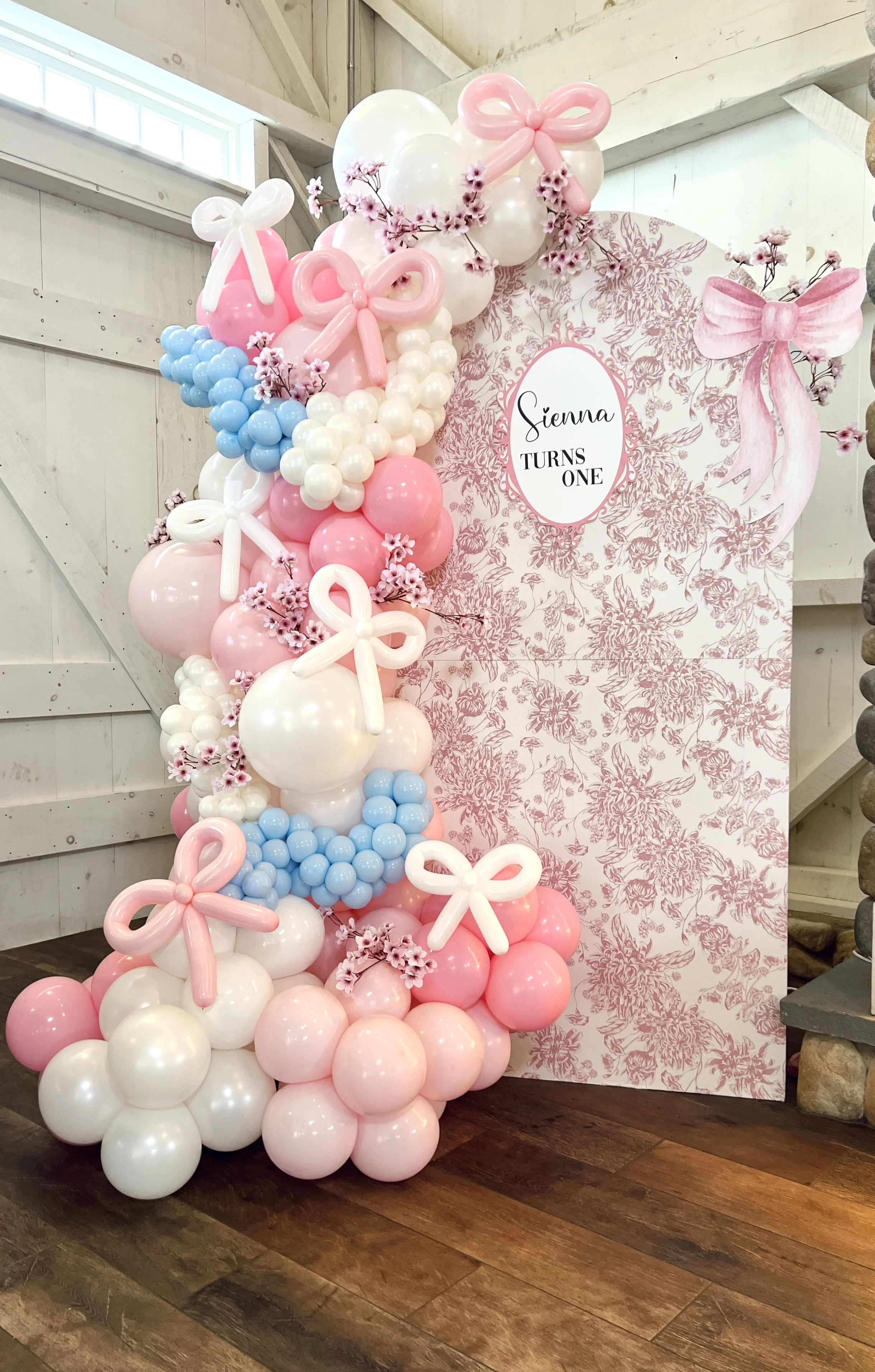 Decorative balloon arrangement in pink, white, and blue balloons with pink bows and flowers next to a pink and white floral themed backdrop with the text 'Sienna Turns One' for a first birthday celebration.