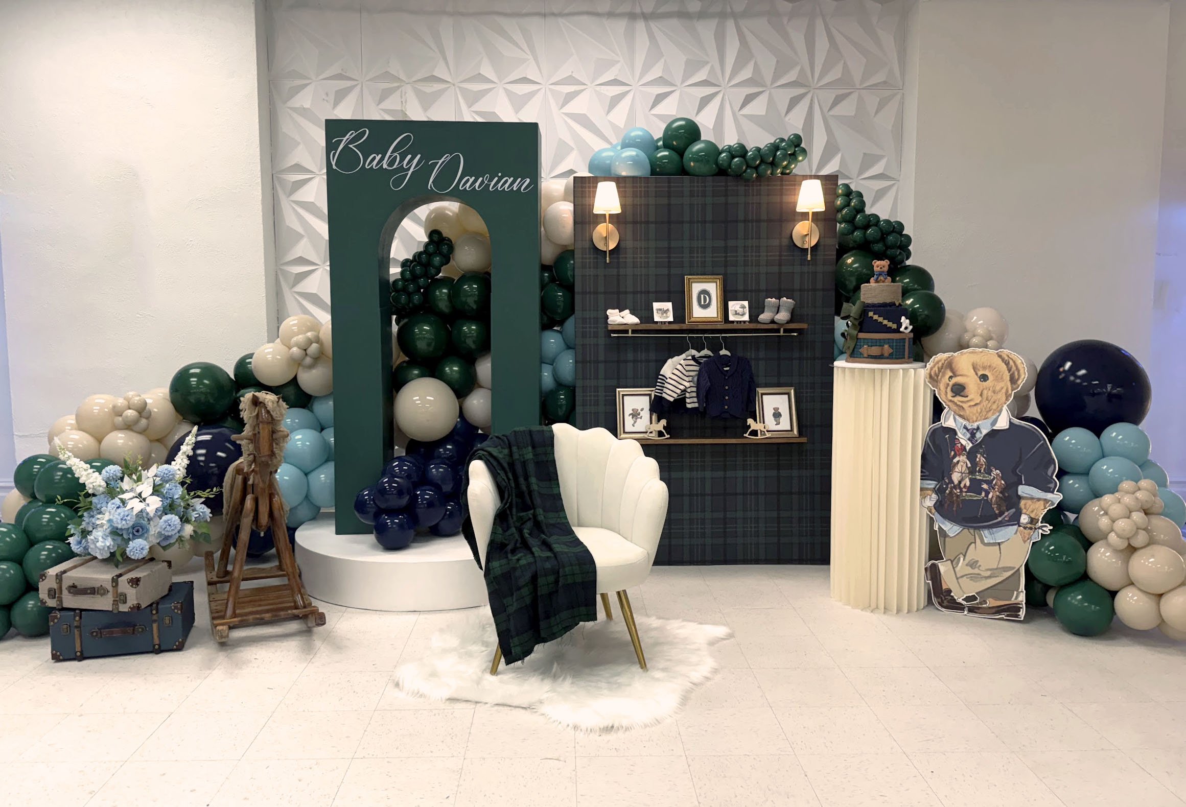 Decorated backdrop for a baby shower or birth celebration with a green wooden structure labeled "Baby Davian," surrounded by green, white, and blue balloons, a white chair with a green plaid blanket, a floral arrangement, and teddy bears.