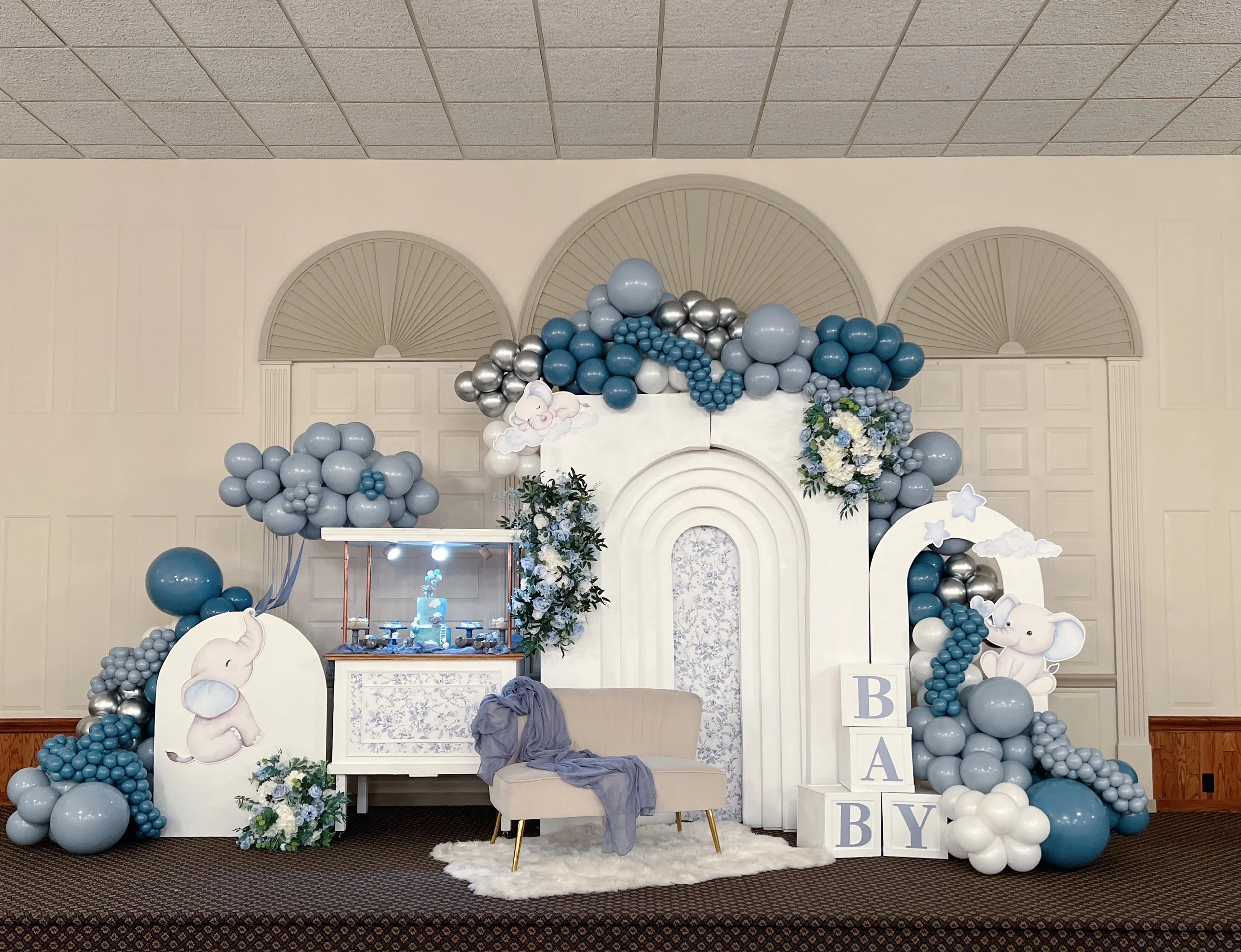 A baby shower backdrop decorated with blue, white, and silver balloons arranged with clouds, flowers, and cartoon elephants. There are painted elephants holding balloons, a white piano, a loveseat with a blue blanket, and decorative blocks spelling '