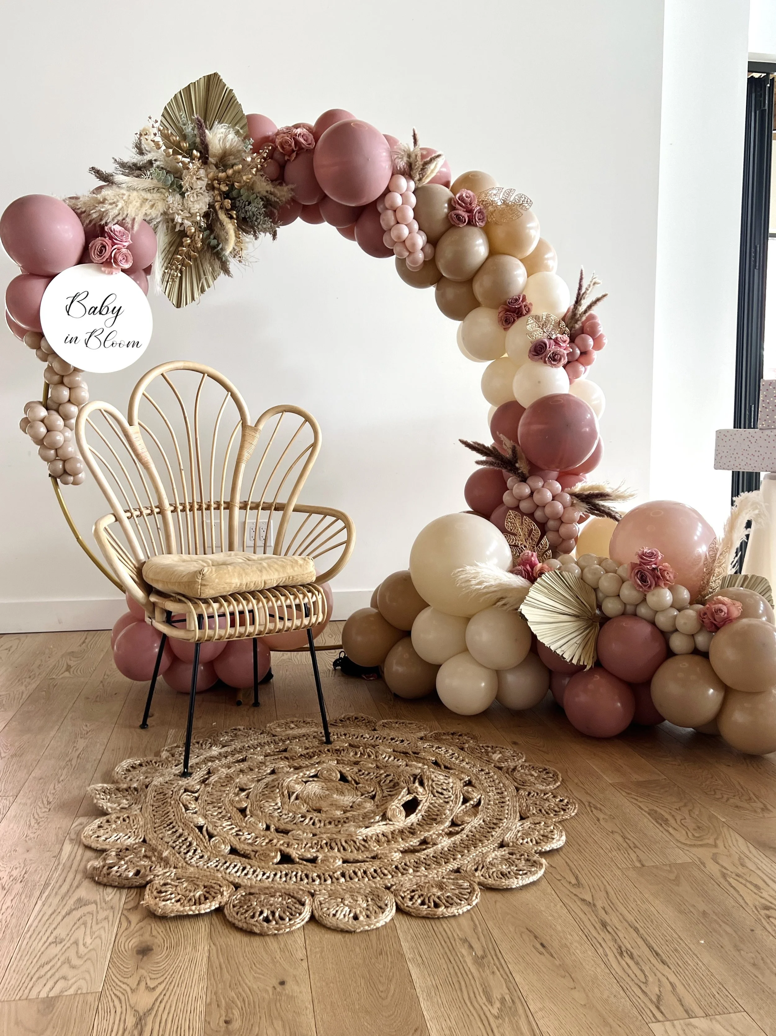 Decorative balloon arch with pink, beige, and white balloons, some with flowers, leaves, and feathers, with a sign that reads "Baby in Bloom" next to a woven chair and a round woven rug on a wooden floor.