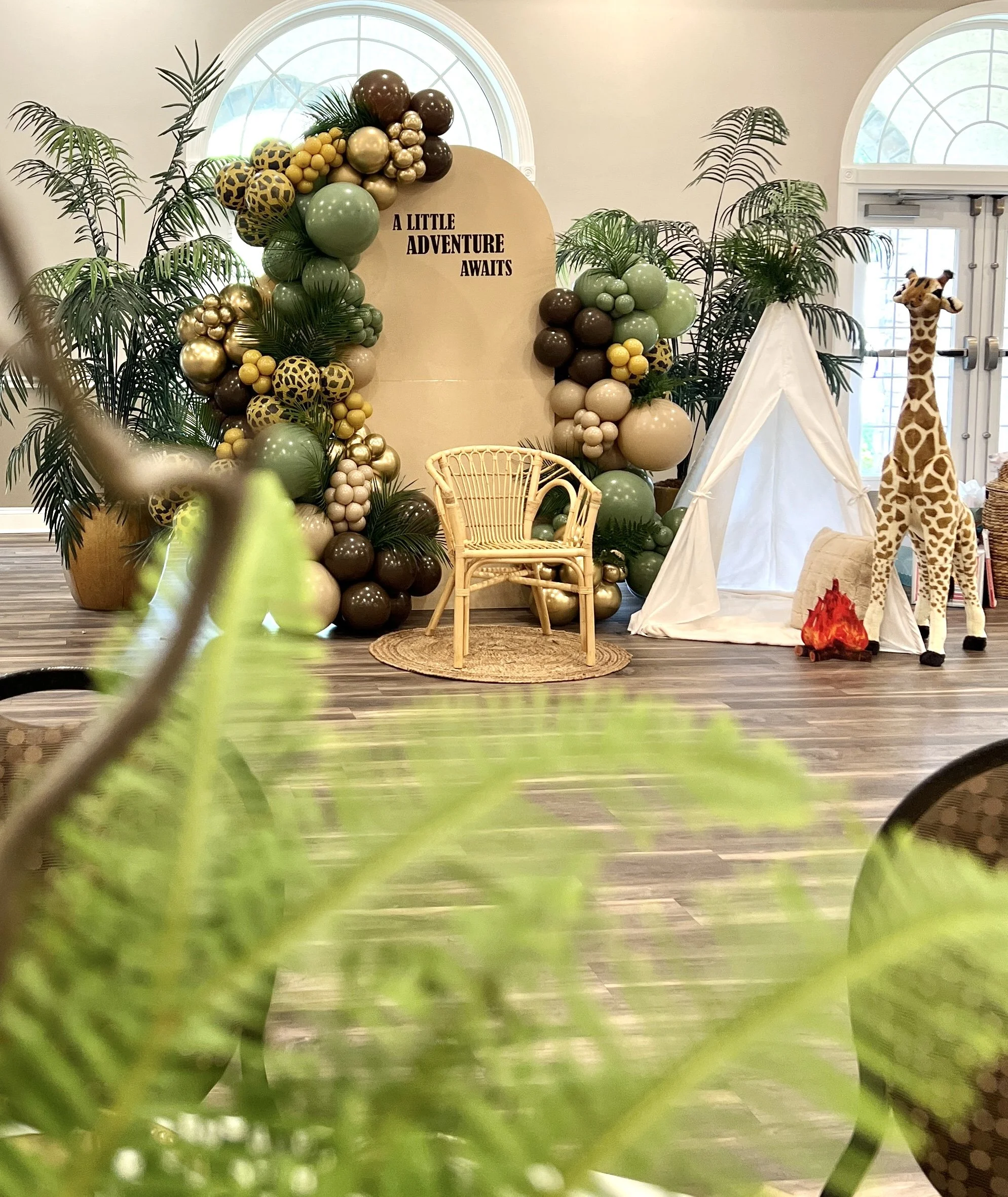 Decorative jungle-themed scene with balloons, a small tent, a giraffe plush toy, and a sign that says 'A Little Adventure Awaits'.