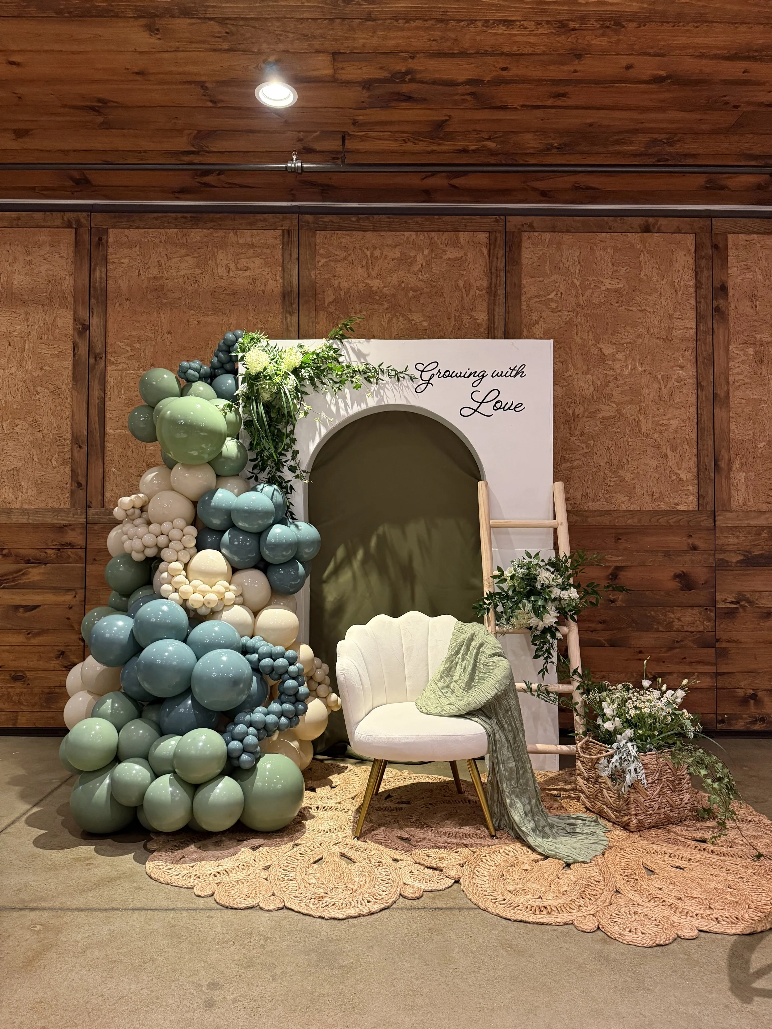 Decorative setup with a balloon arrangement, a white chair with a green cloth, a floral arrangement, and a backdrop with the words "Growing with Love" in elegant script, inside a room with wooden walls and ceiling.