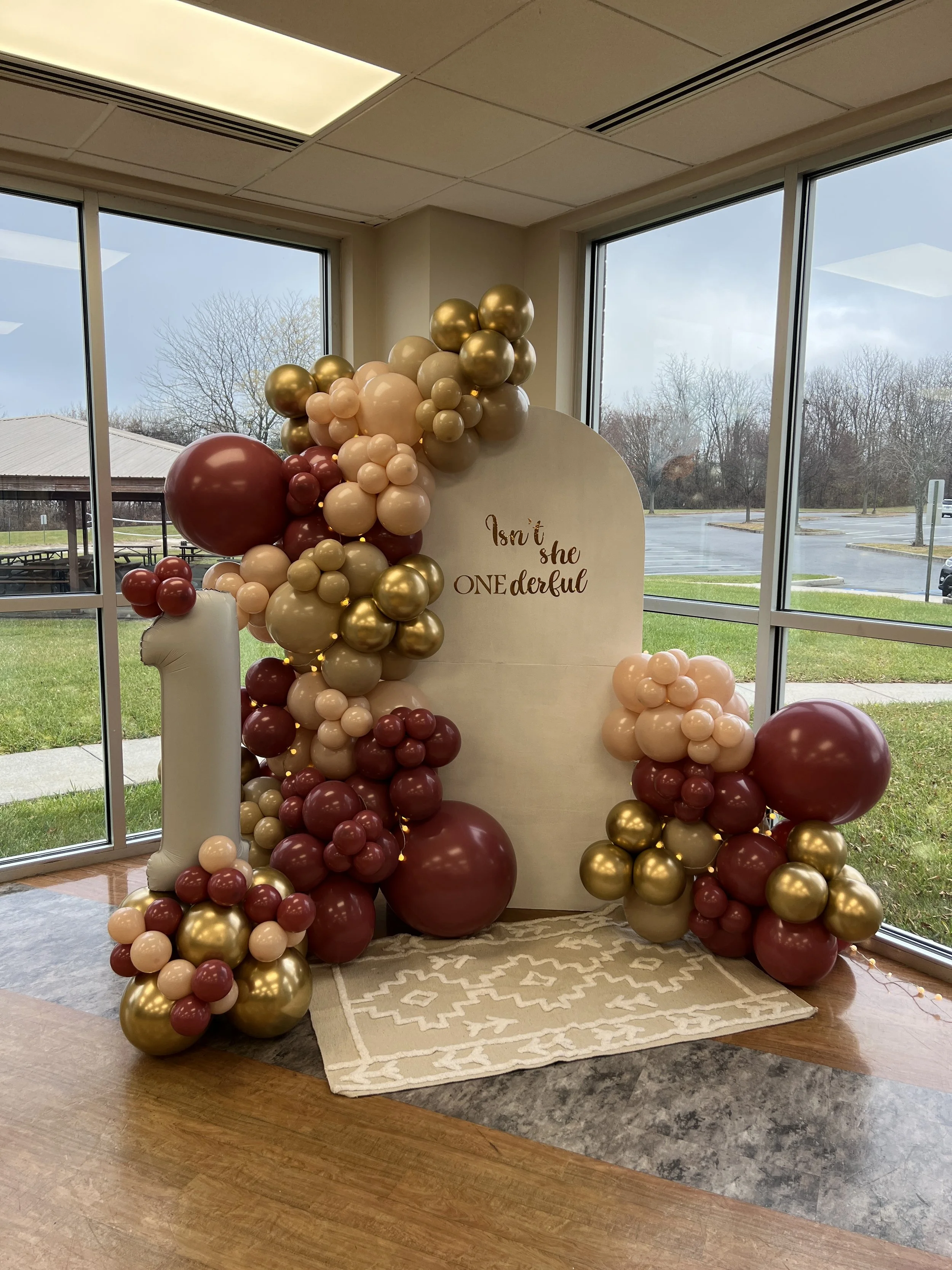 Balloon decoration with a white background, large white number one, and a gold, beige, and burgundy color theme, with the text "Isn't she ONE derful" on the background for a first birthday celebration.