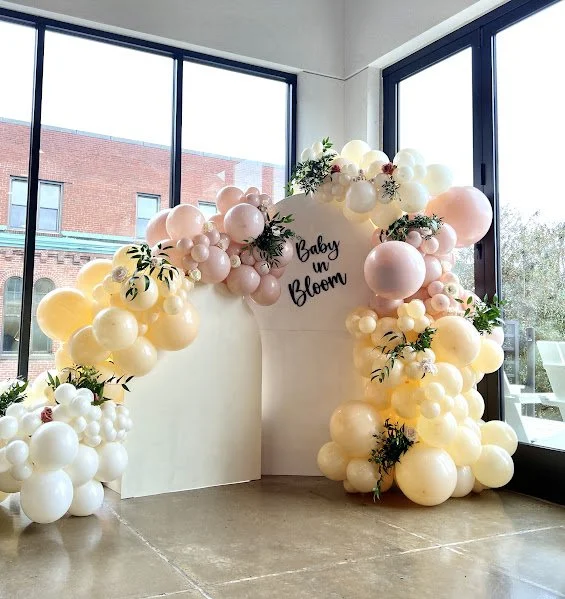 Decorative arch made of pink, white, and cream balloons with greenery, set inside a glass-enclosed space with large windows, celebrating a baby shower named 'Baby on Bloom'.