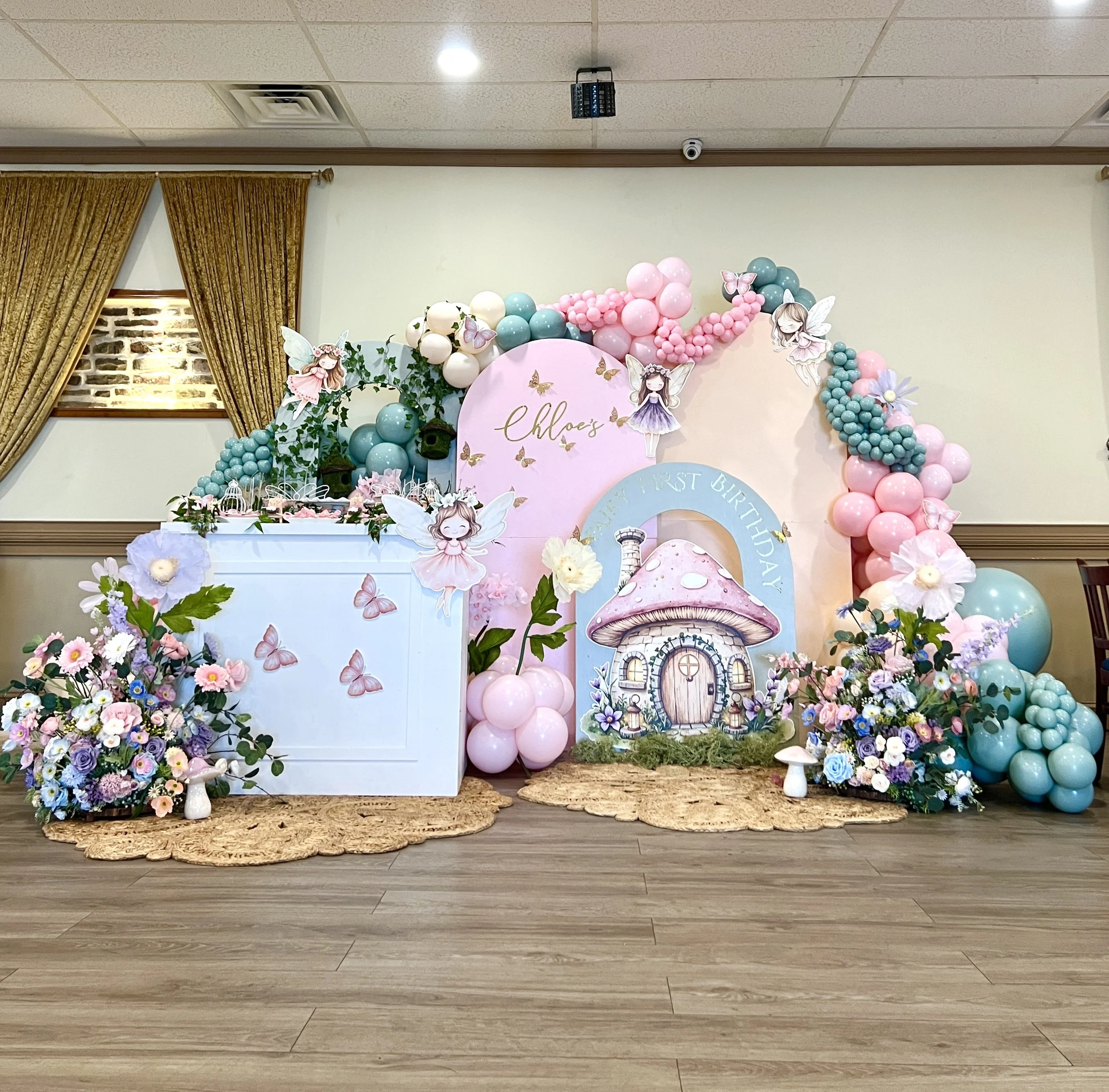 Decorated area for a child's first birthday party featuring fairy tale theme with pink and blue balloon arrangements, floral arrangements, fairy figurines and a whimsical house illustration.