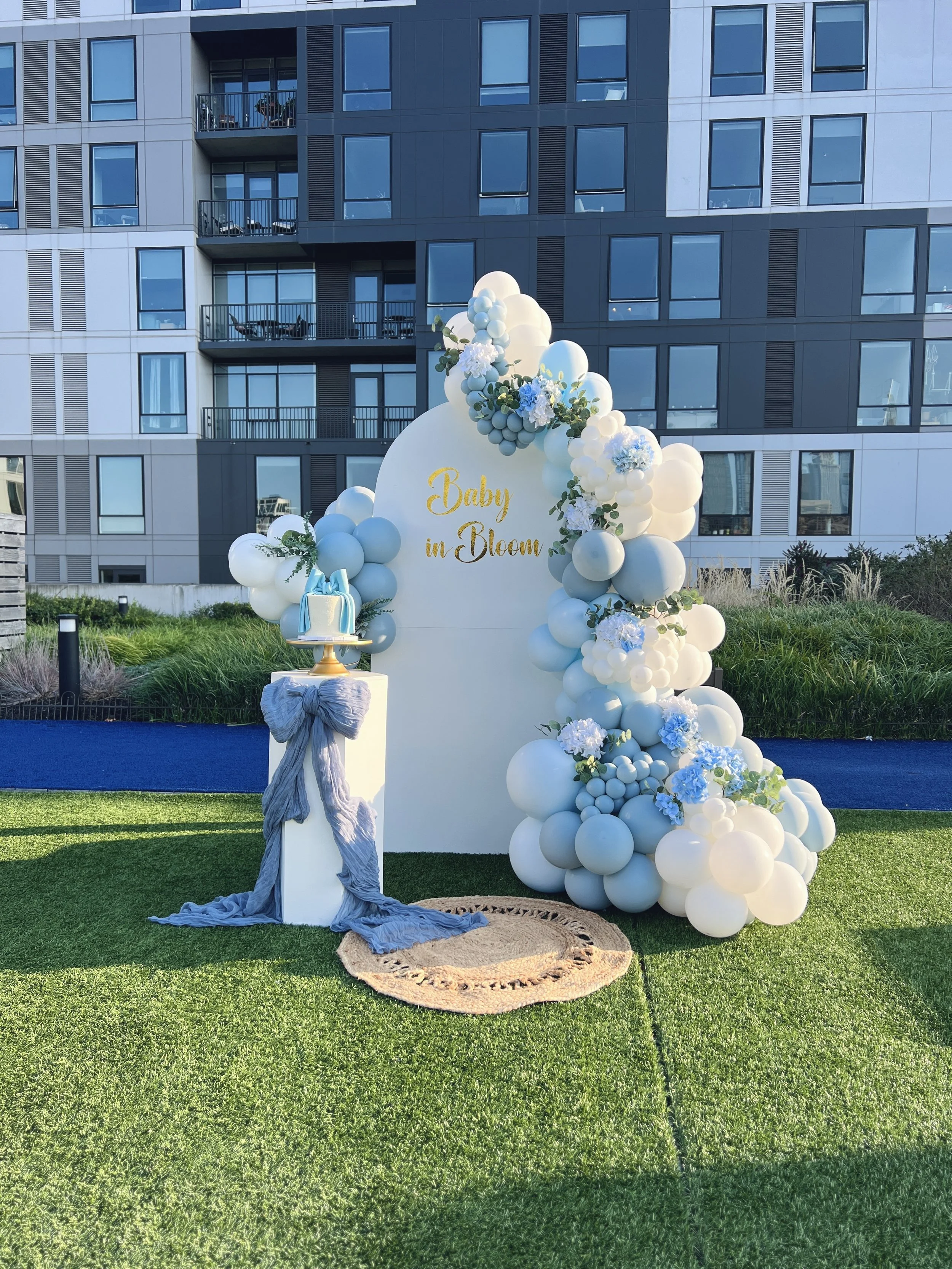 Decorative outdoor setup with a white arch decorated with blue and white balloons, green foliage, and white flowers, with a sign that reads 'Baby in Bloom.' A small platform with a cake and a blue ribbon are next to the arch, on a grass lawn with a m