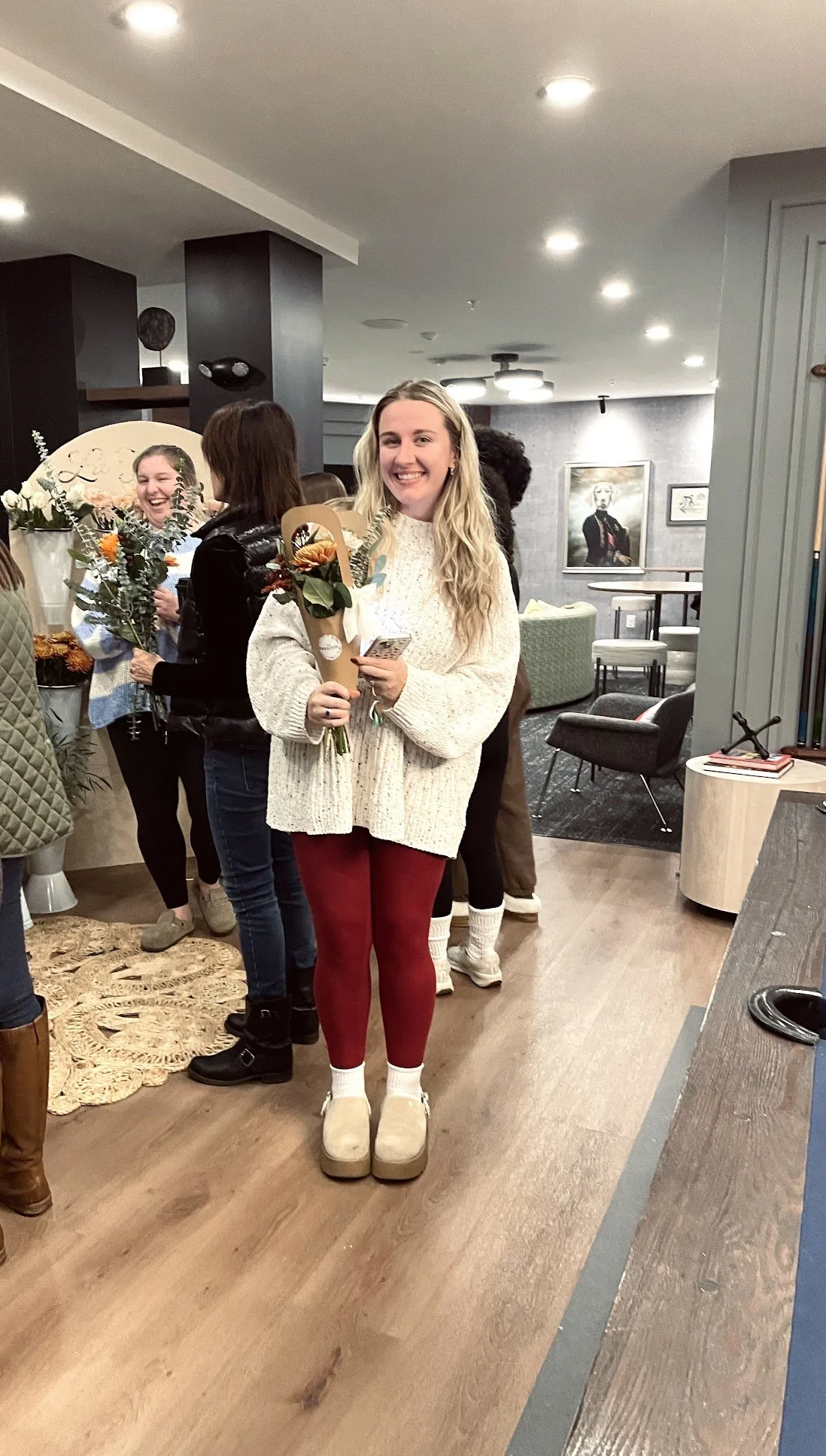 A young woman holding a bouquet of flowers and a card, smiling at the camera in a room with several people in the background.