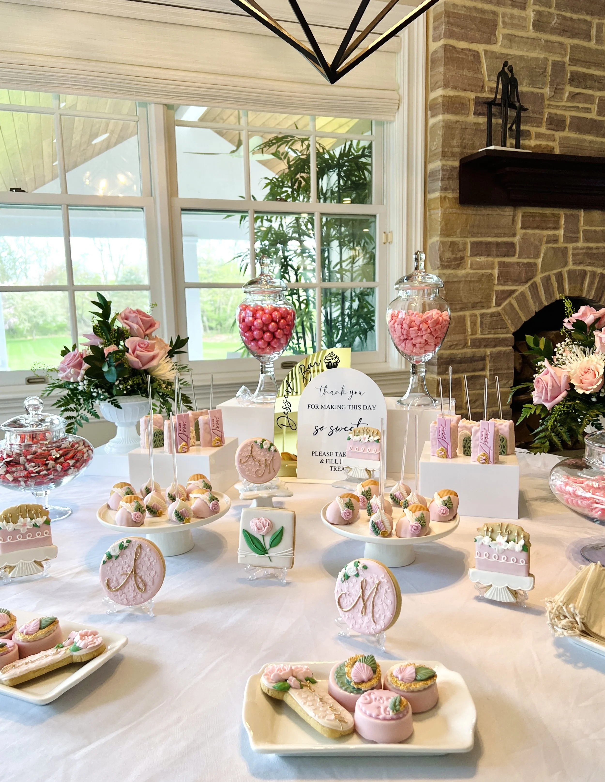 A dessert table decorated with pink and white treats, including cookies, cake pops, and candies, with floral arrangements and a thank you sign in a bright room with large windows and brick walls.