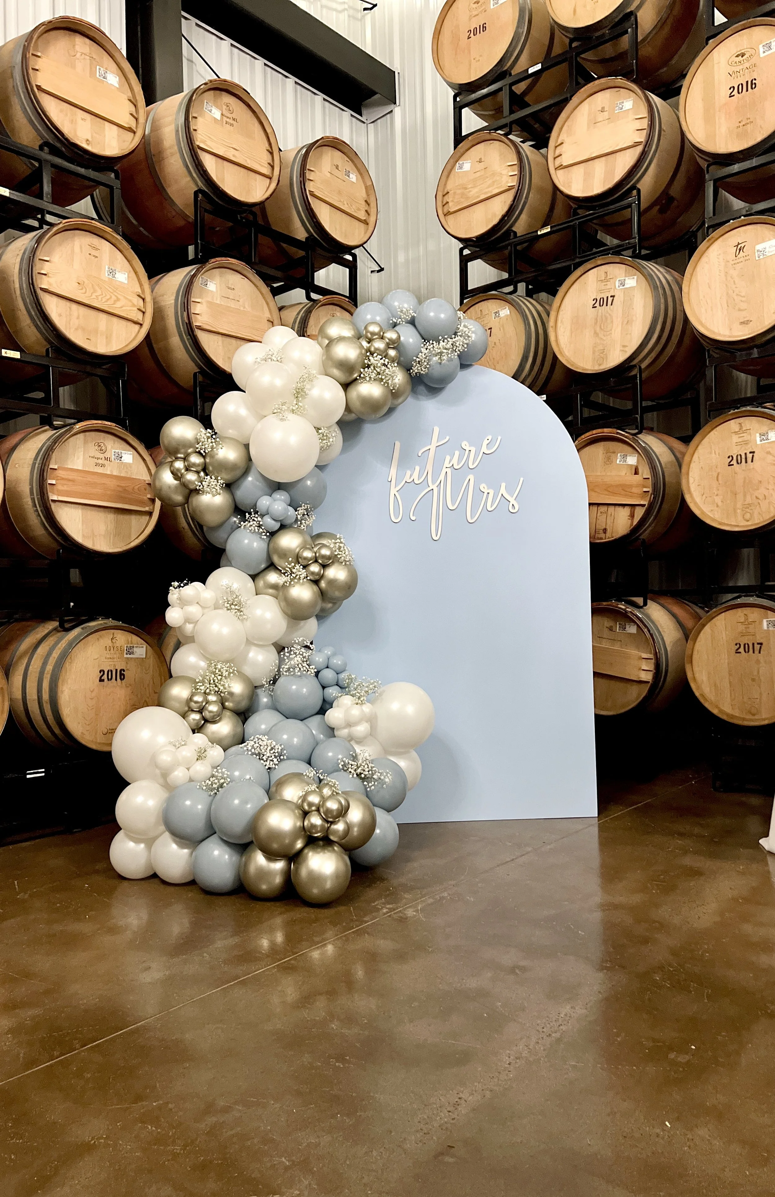 Decorative balloon arrangement with white, silver, and grey balloons next to a blue display board labeled 'future Mrs' in a room with wine barrels in the background.