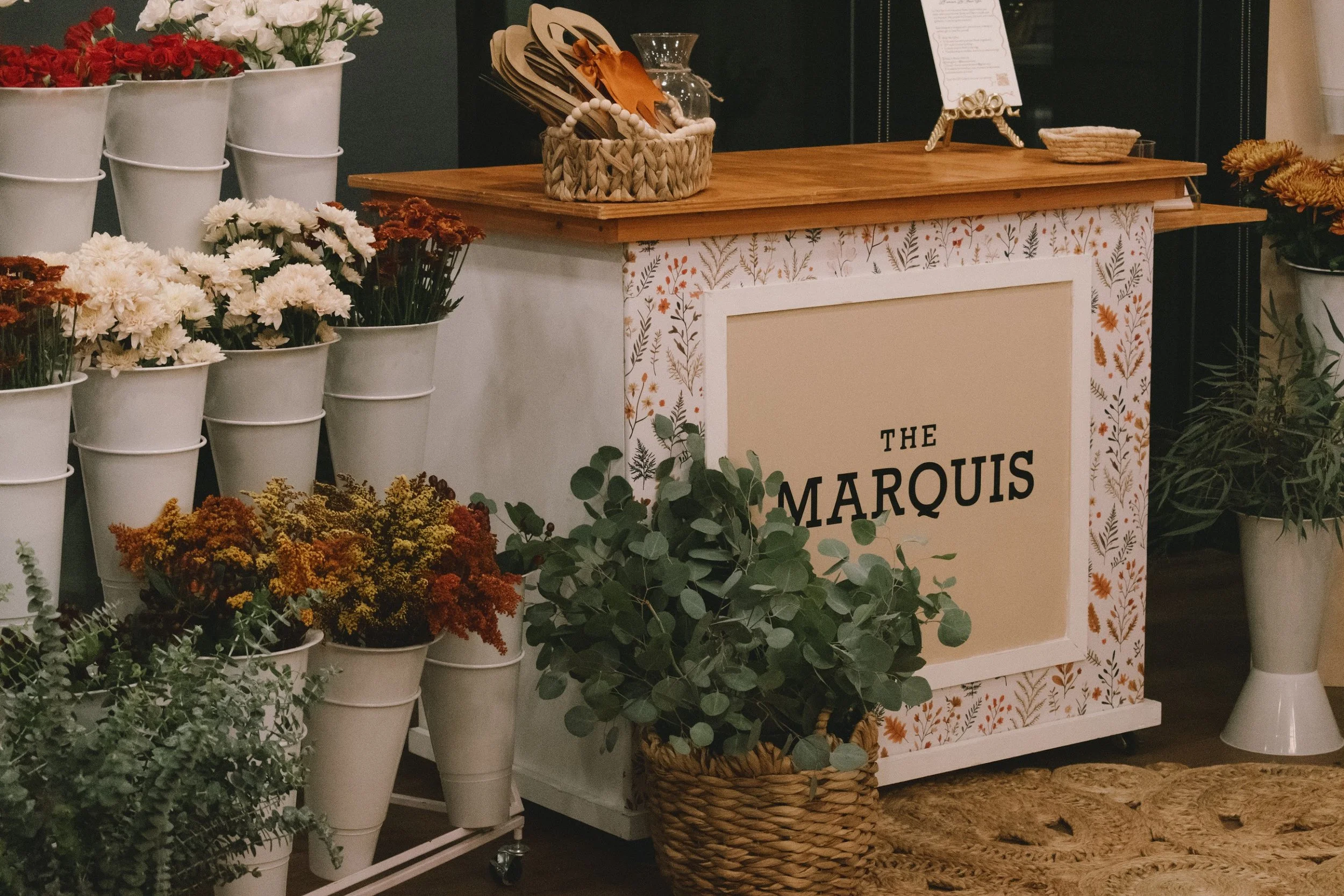 A flower shop counter with white buckets of white, red, orange, and yellow flowers, a wicker basket with green foliage, and a decorative sign reading 'The Marquis' decorated with floral patterns.