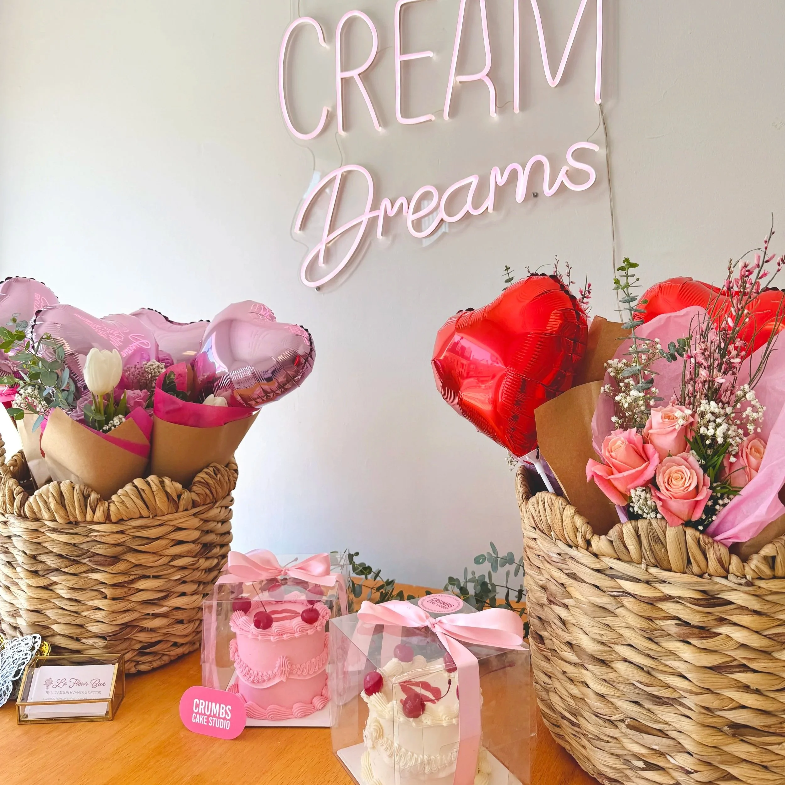 Decorative pink and red heart-shaped balloons, two baskets filled with pink roses and other flowers, pink and white cakes with cherries, and a neon sign reading 'CREAM Dreams' on a plain wall.