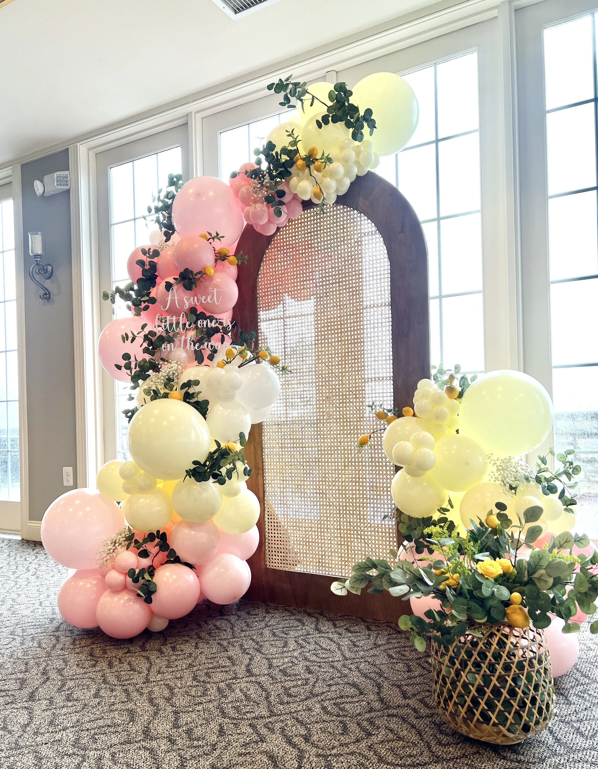Decorative balloon arrangement with pink, yellow, and white balloons, greenery, and flowers surrounding a wooden door with a mesh panel, in front of large windows.