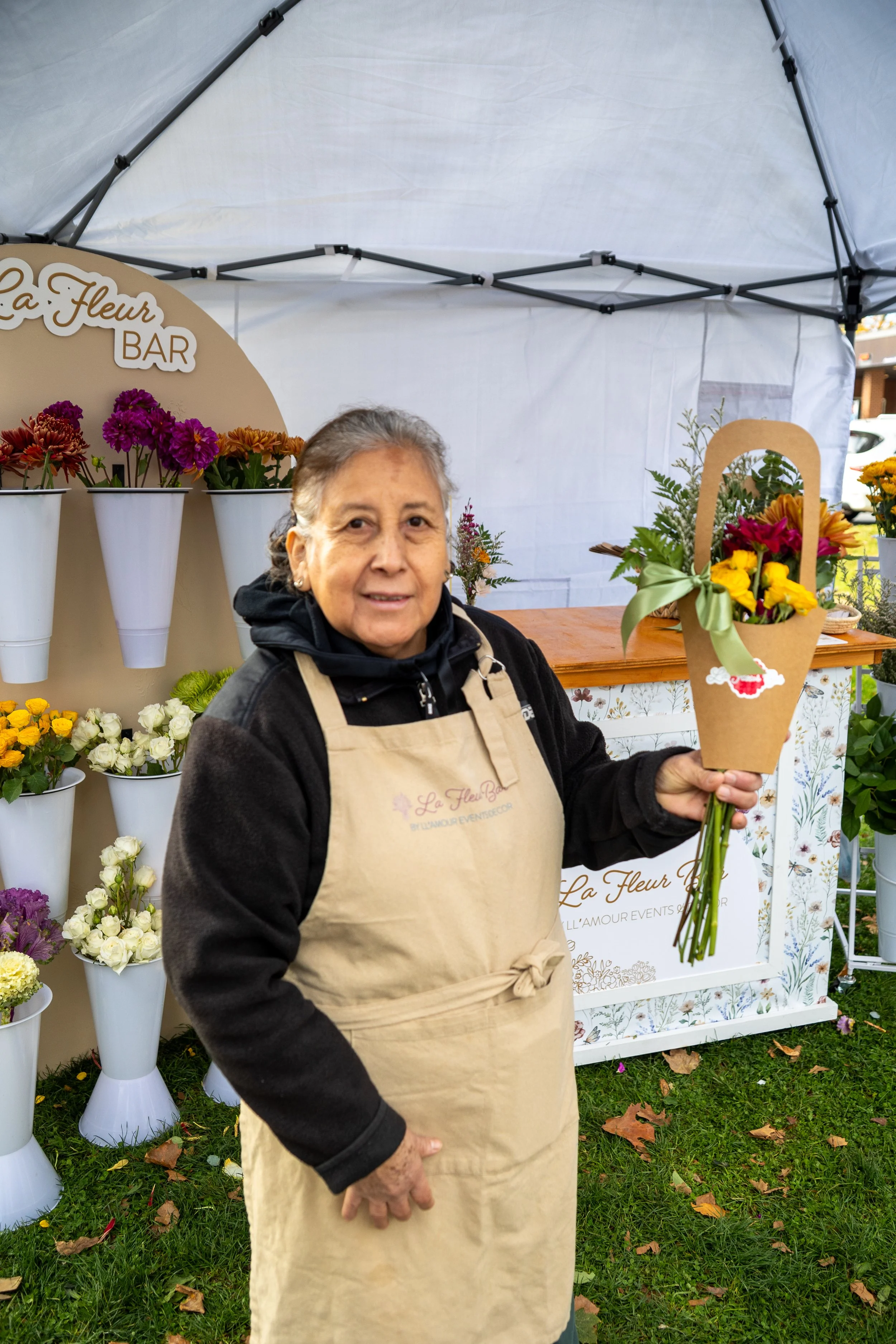 A woman standing in front of a flower stand holding a bouquet of flowers in a brown paper bag. The stand has a sign that says ‘La Fleur BAR’ and displays colorful flowers in white vases.