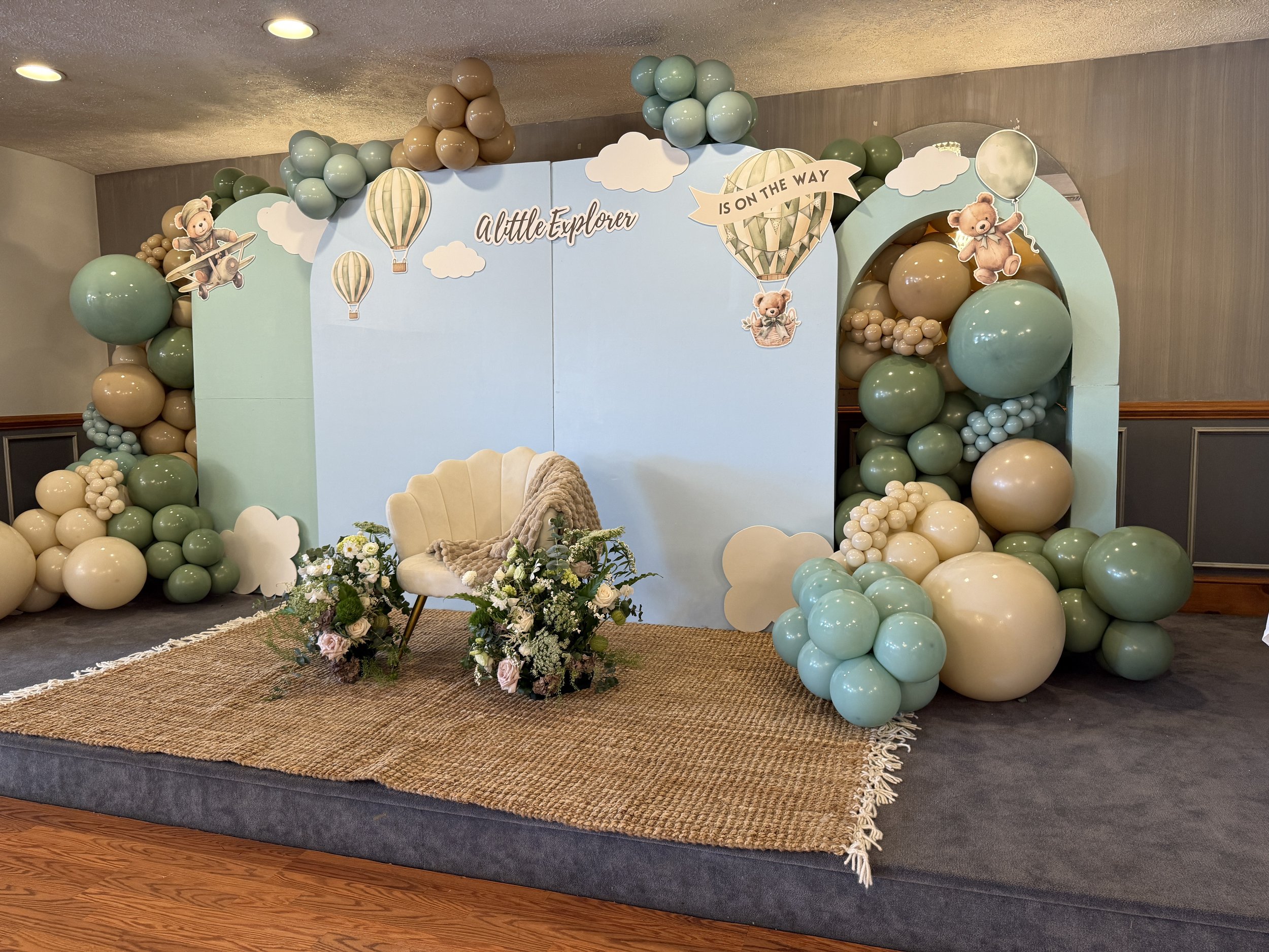 Decorative backdrop for a child's birthday or celebration featuring balloons in pastel greens, blues, and beige, with cartoon teddy bear and animal hot air balloon cutouts, a white tufted chair adorned with floral arrangements, and a blue sky-themed 