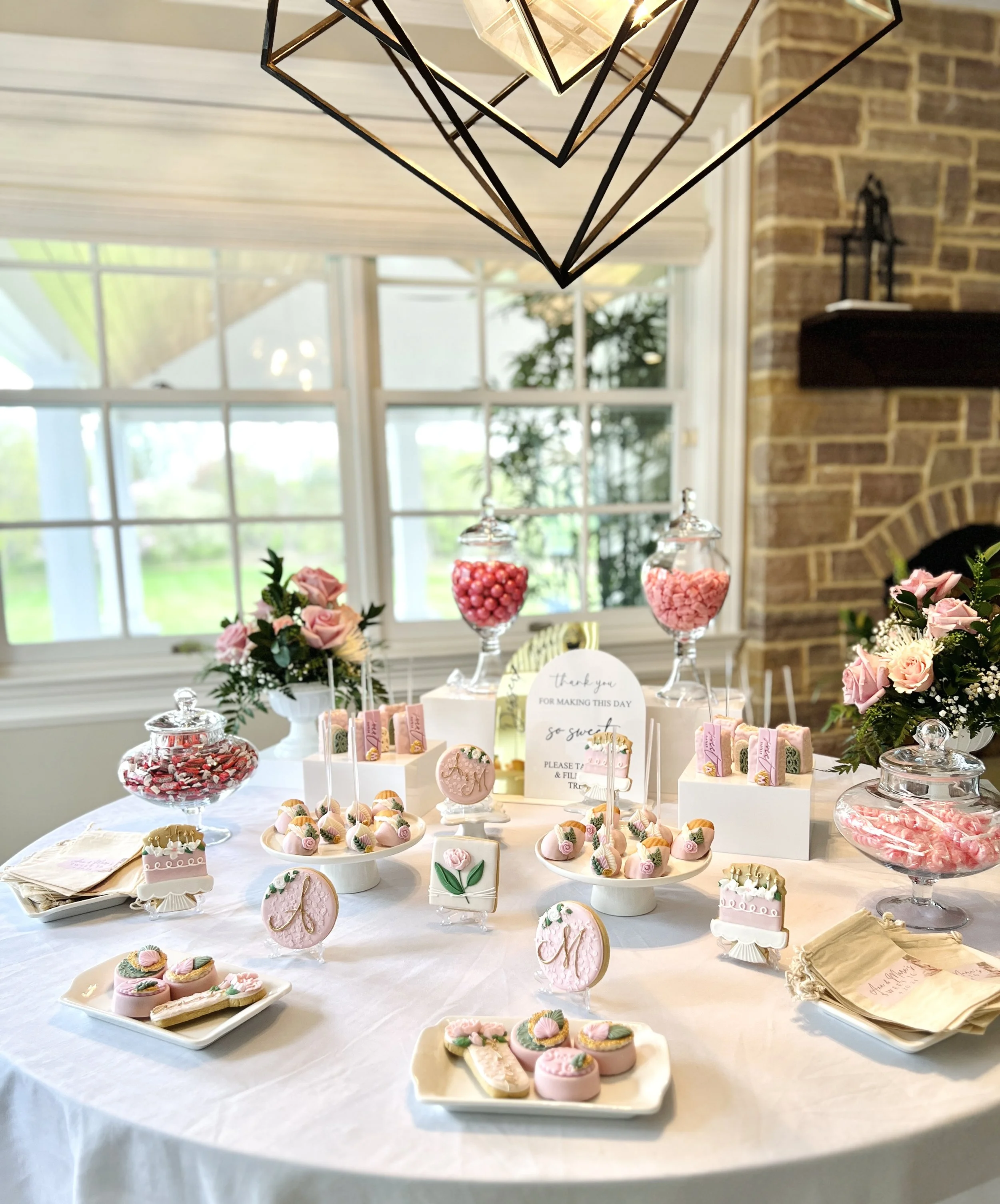 A dessert table decorated with pink sweets, flowers, and a thank you note, set against a backdrop of a large window and brick wall, with a modern black geometric chandelier hanging above.