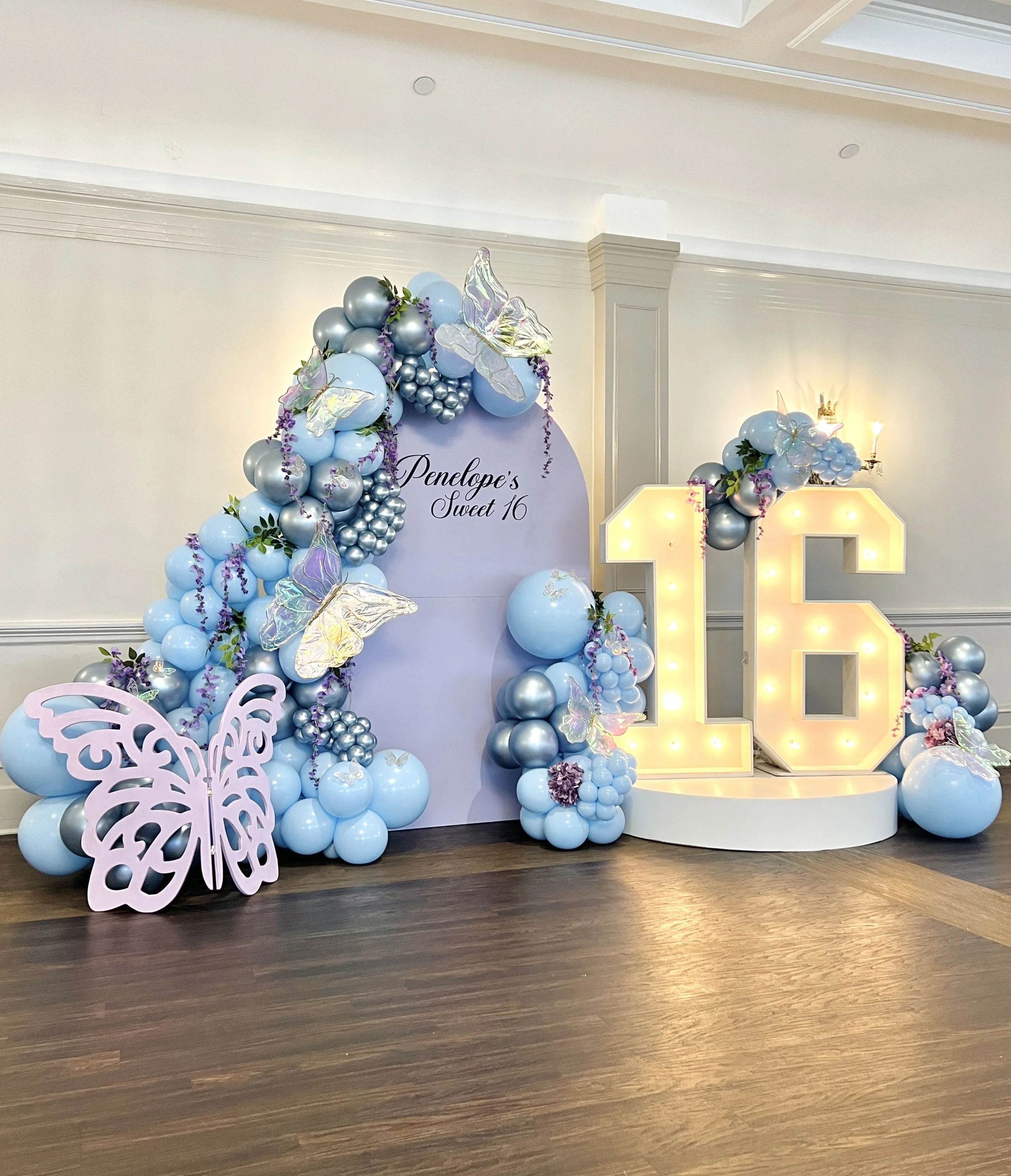 Decorative birthday setup with blue and silver balloons, butterflies, flowers, and a large illuminated number 16. A backdrop reads "Penelope's Sweet 16."