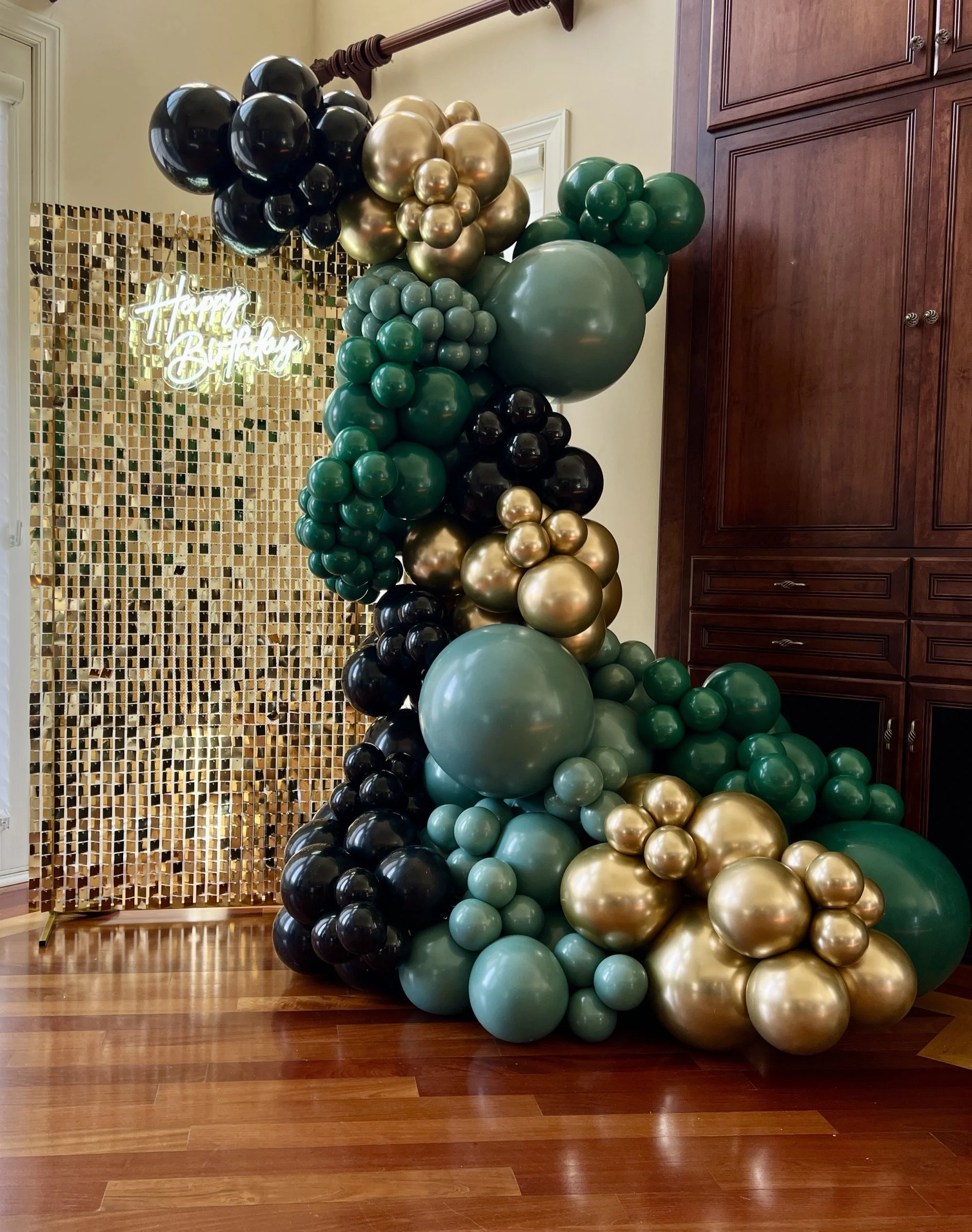 Colorful balloon arrangement with black, gold, and green balloons, and a neon sign that says "Happy Birthday" in the background.