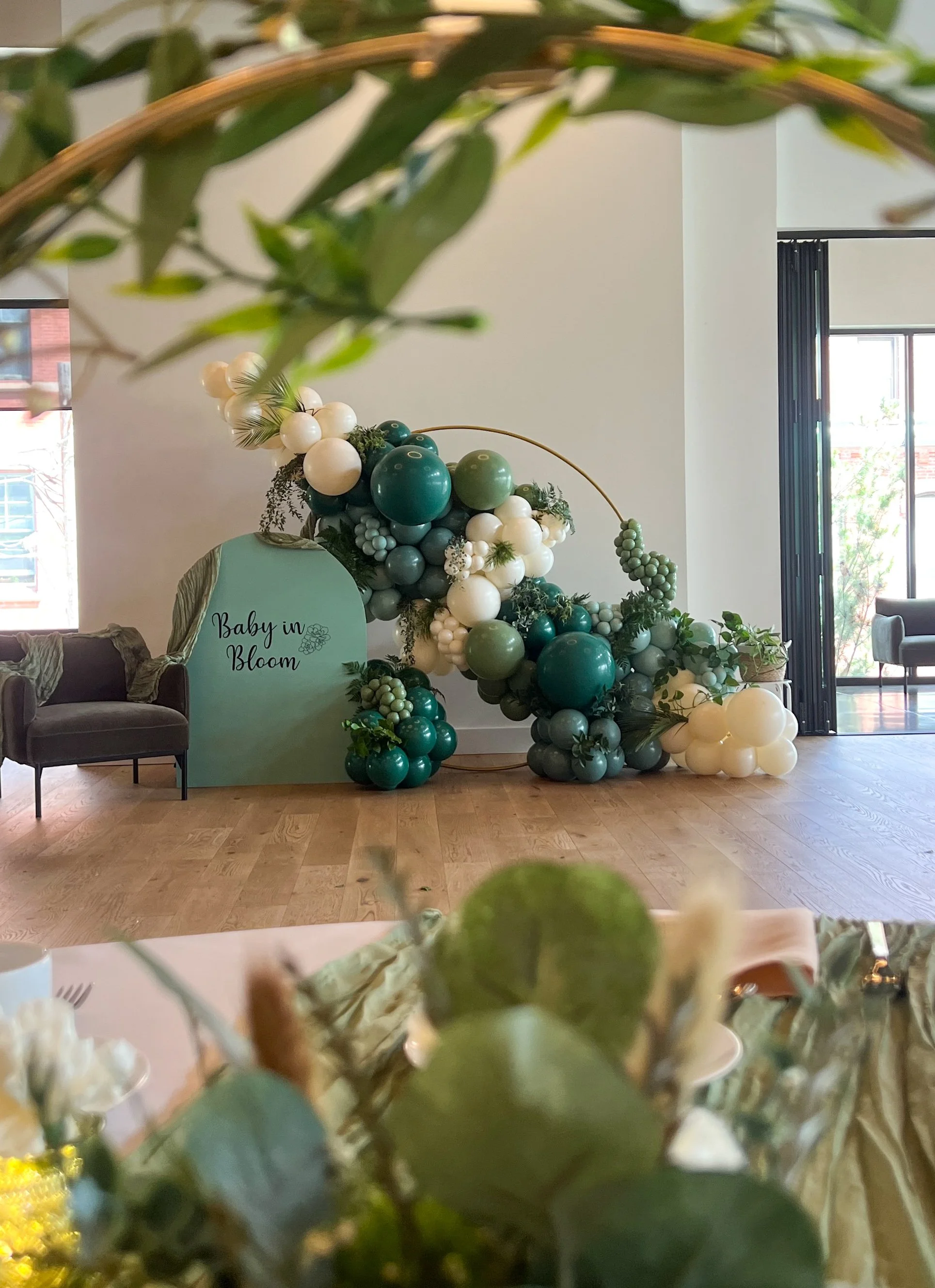 Balloon decoration with a sign that reads "Baby in Bloom" for a baby shower, featuring green, white, and teal balloons arranged with greenery, set inside a bright, modern room with wooden flooring, furniture, and large windows.