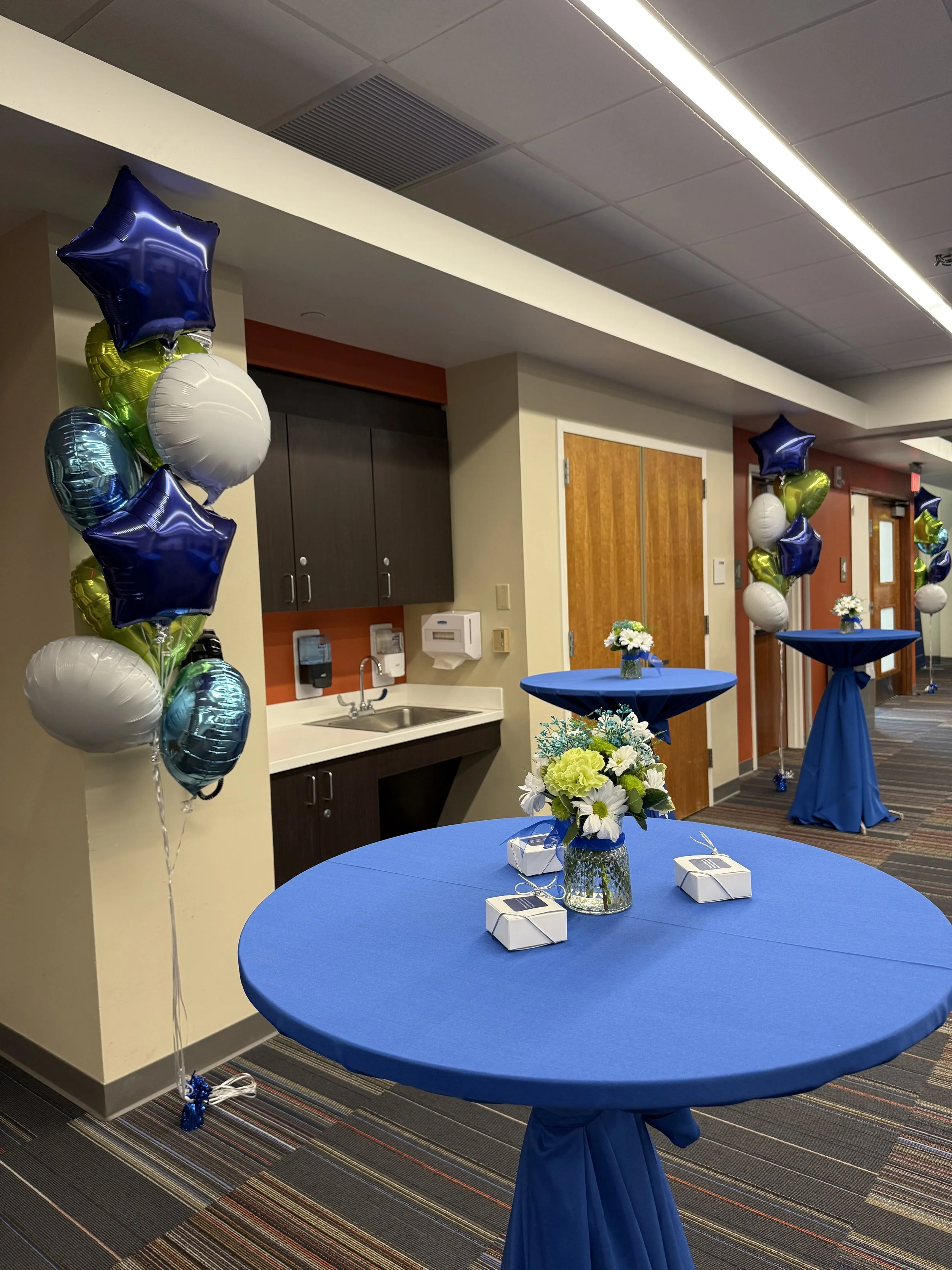 Decorated event space with blue tablecloths, floral centerpieces, balloon columns, and gift boxes.