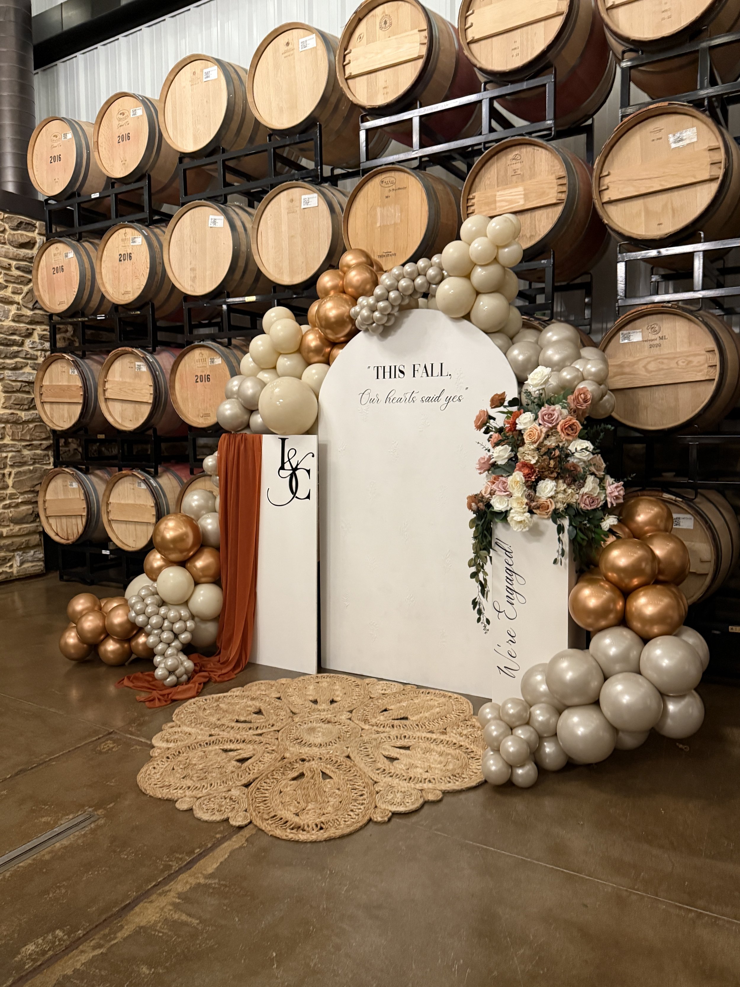 Decorative wedding or event backdrop with balloons, flowers, and a sign that reads 'This fall, our hearts said yes. You're engaged!' in front of a winery or brewery with stacked wine barrels on racks.