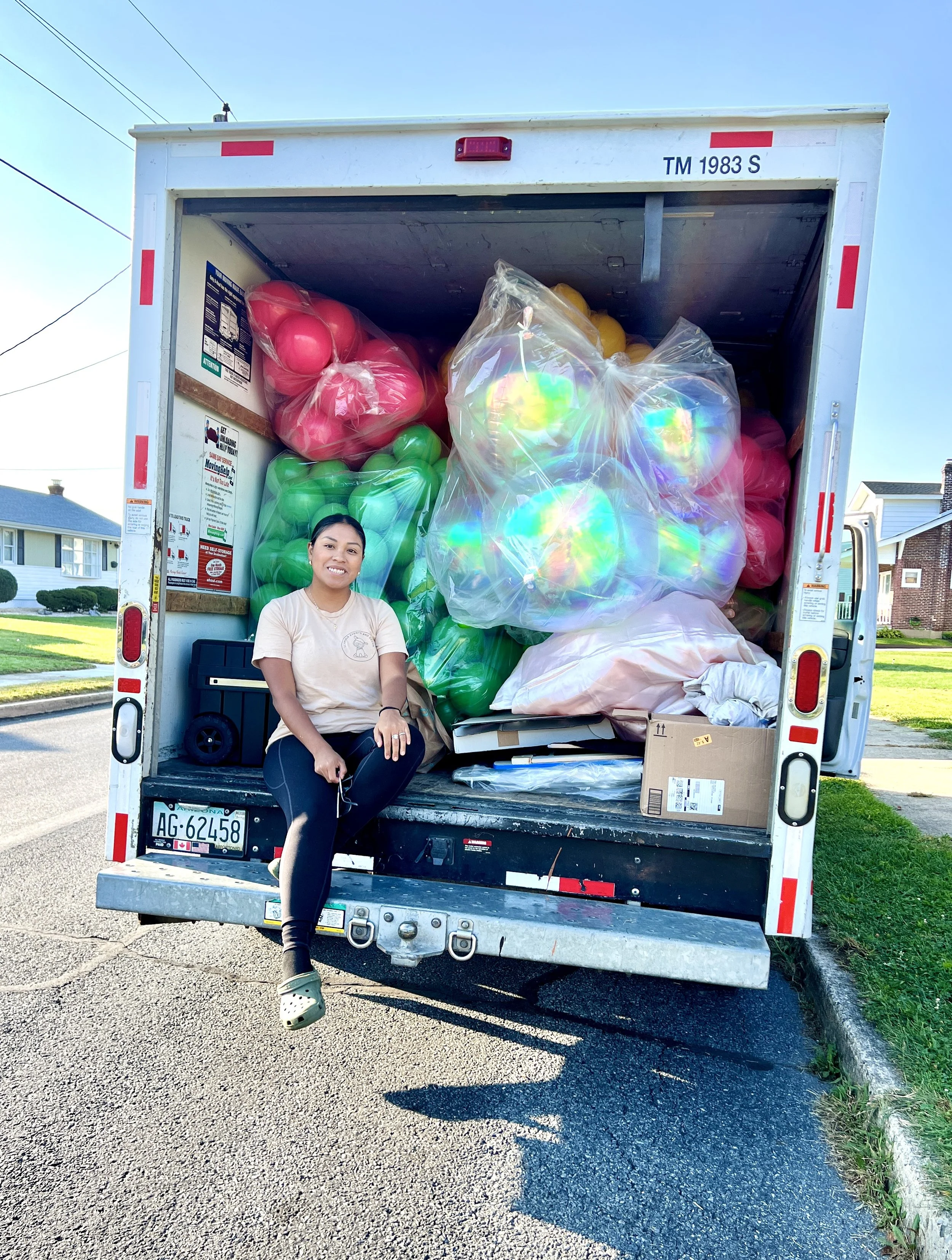 A woman sitting on the back of a truck filled with large, colorful balloons, including pink, green, and iridescent ones, parked on a residential street.