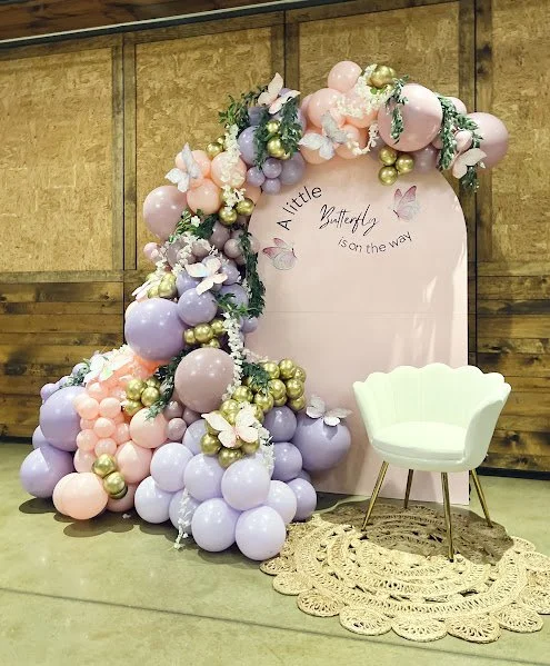 Decorative backdrop with pastel-colored balloons, flowers, and a pink sign reading 'A Little Butterfly is on the way,' accompanied by a white shell-shaped chair and a lace rug.