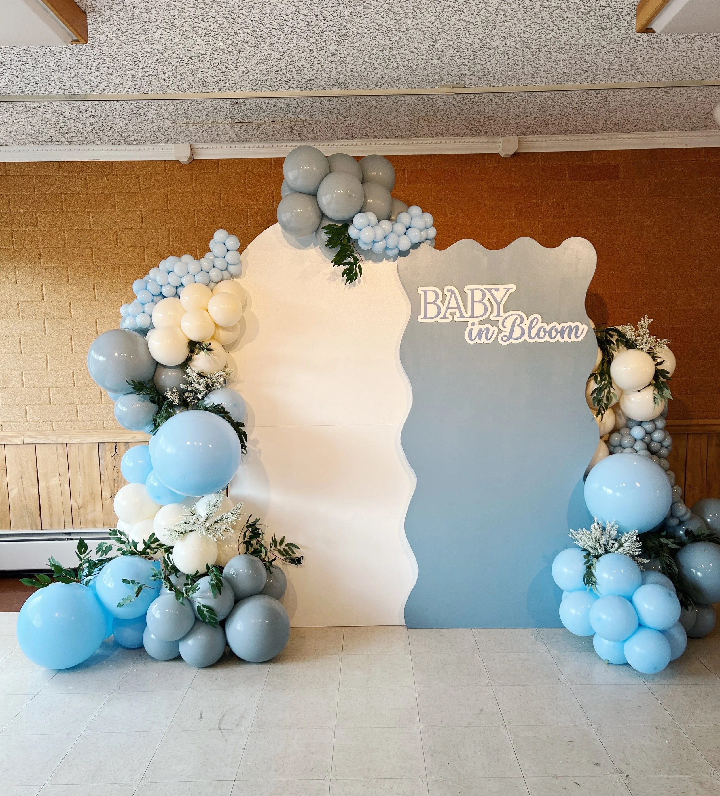 Decorative backdrop with balloon arrangements in shades of blue, white, gray, and greenery, featuring the text "BABY in Bloom" on a wavy board for a baby shower or celebration.