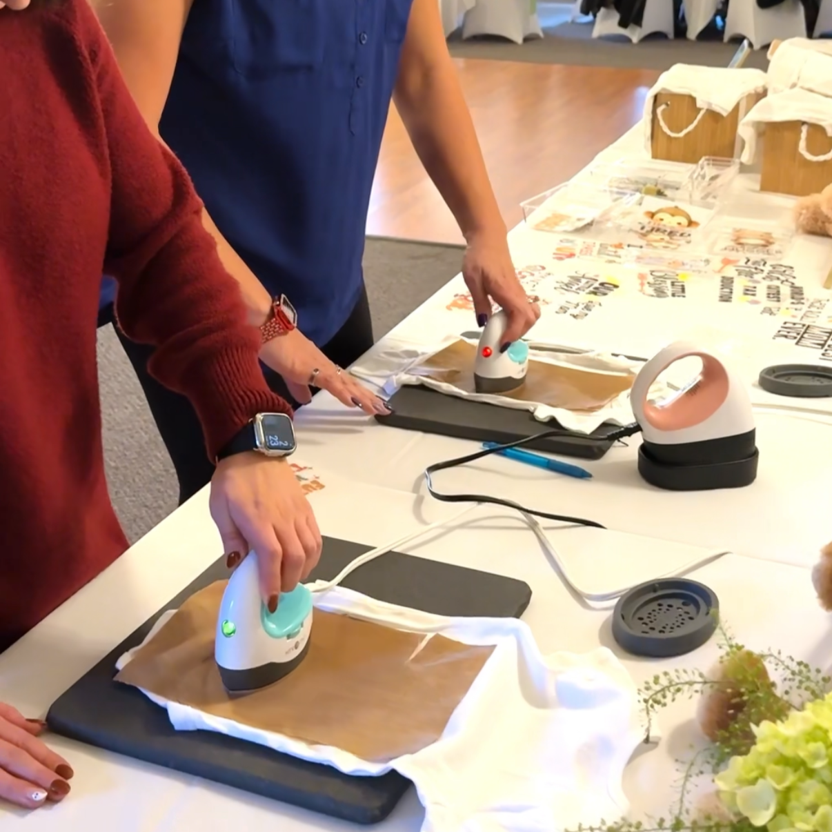People using small handheld irons on fabric-covered boards at a table with printed papers and decorative items nearby.