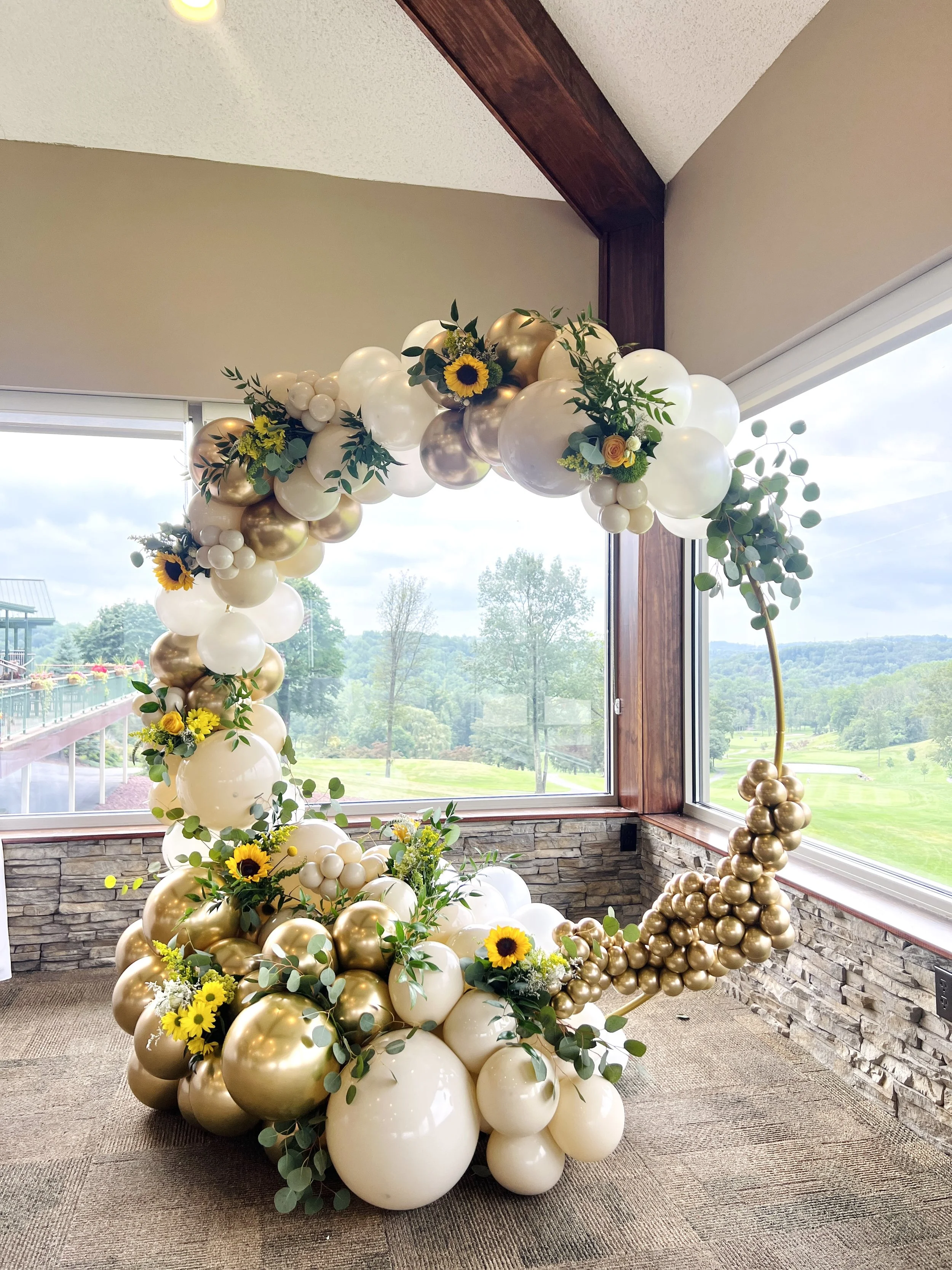 Balloon arch with white, gold, and silver balloons, decorated with sunflowers and greenery, near large windows showing a green outdoor landscape.