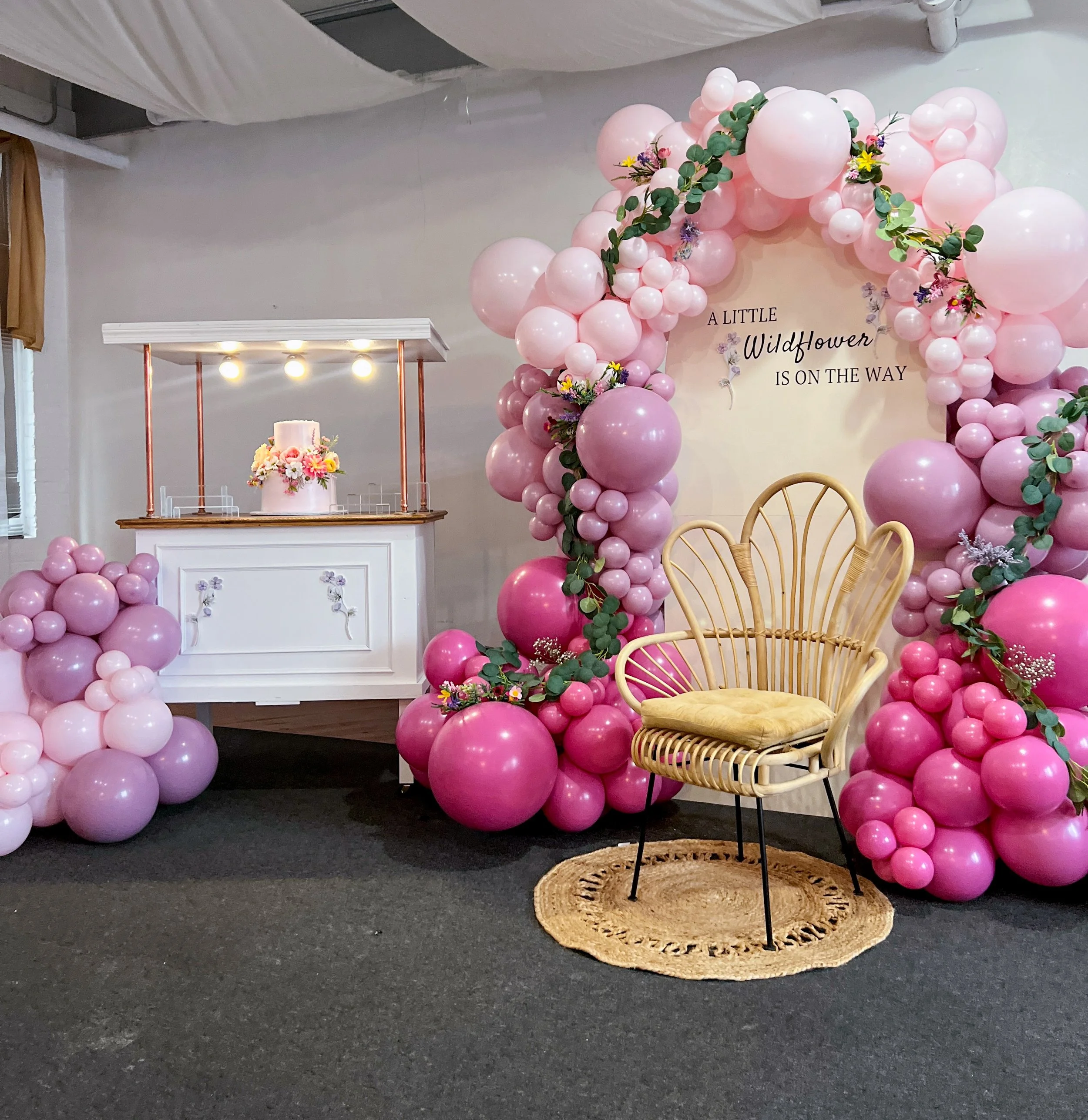 Decorative setup with pink and purple balloons forming an arch and clusters, a white small stand with a floral arrangement and cake, a rattan chair with a yellow cushion, and a backdrop with the text "A little wildflower is on the way."