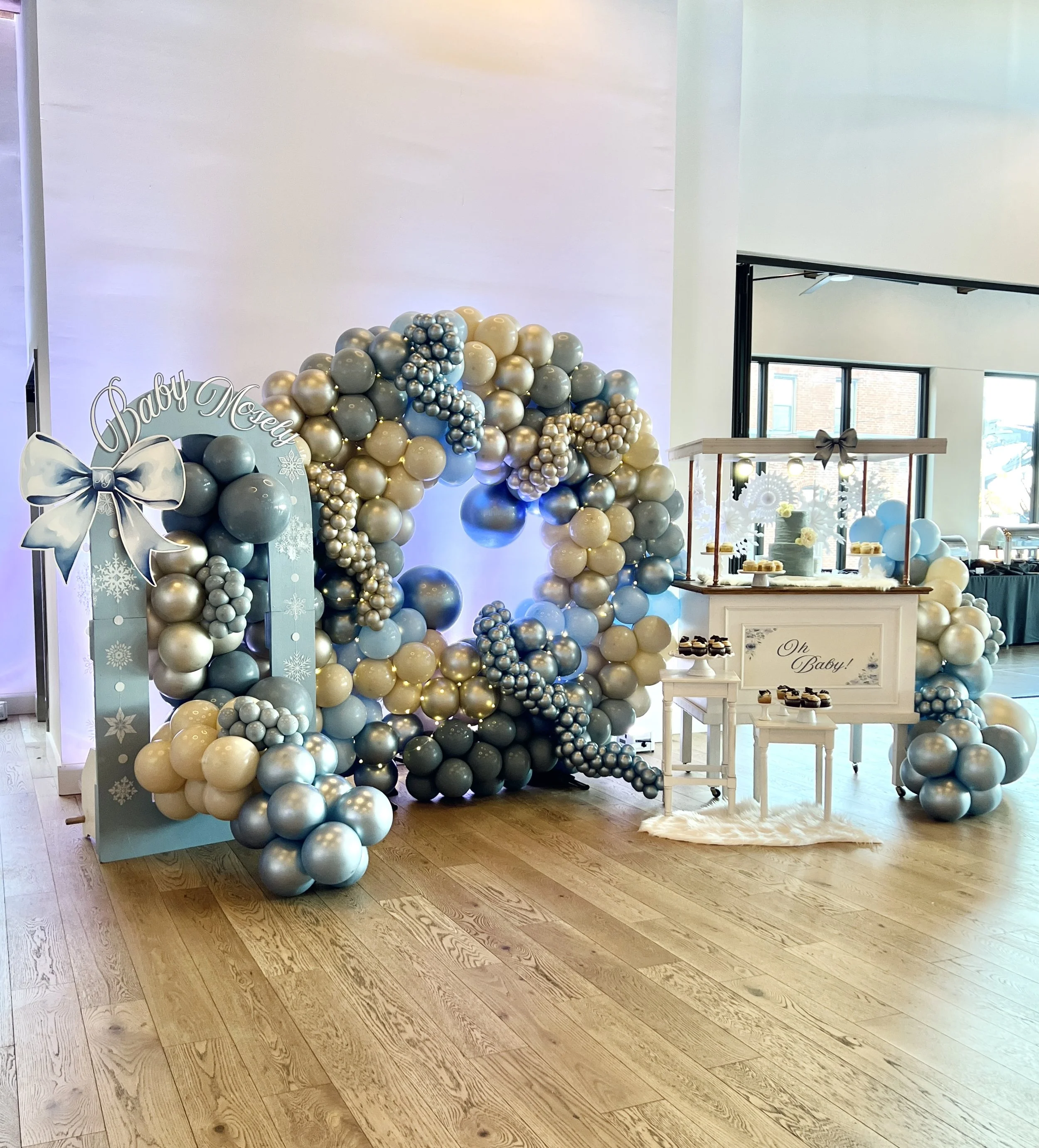 Balloon arrangement in shades of blue, gold, and silver with a decorative frame sign saying "Baby Mochi" and a matching table with cupcakes for a baby shower.