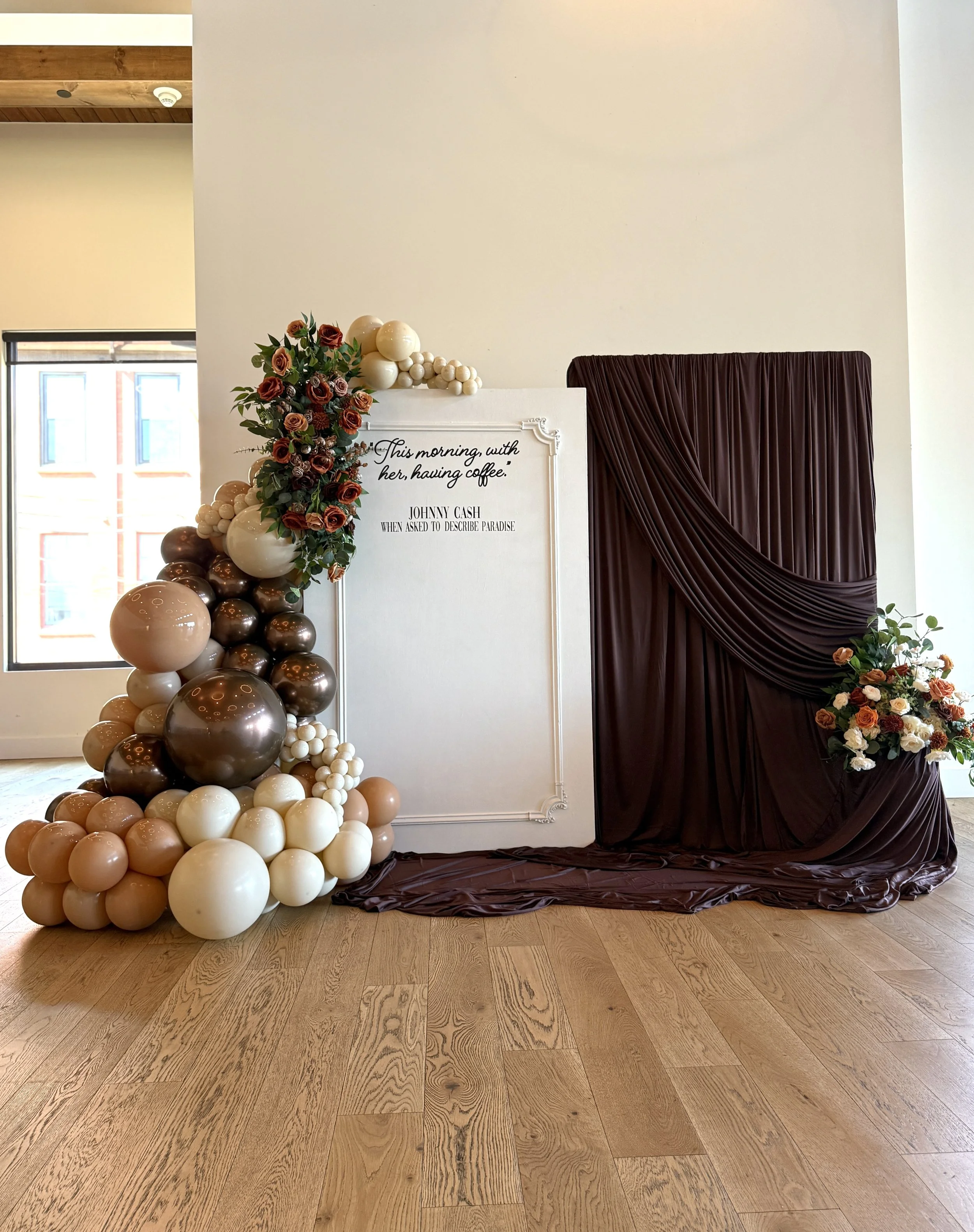 Decorative setup with balloons, flowers, and a sign for an event, featuring a white and brown balloon arrangement and a floral arrangement on a whiteboard, with a red curtain backdrop.