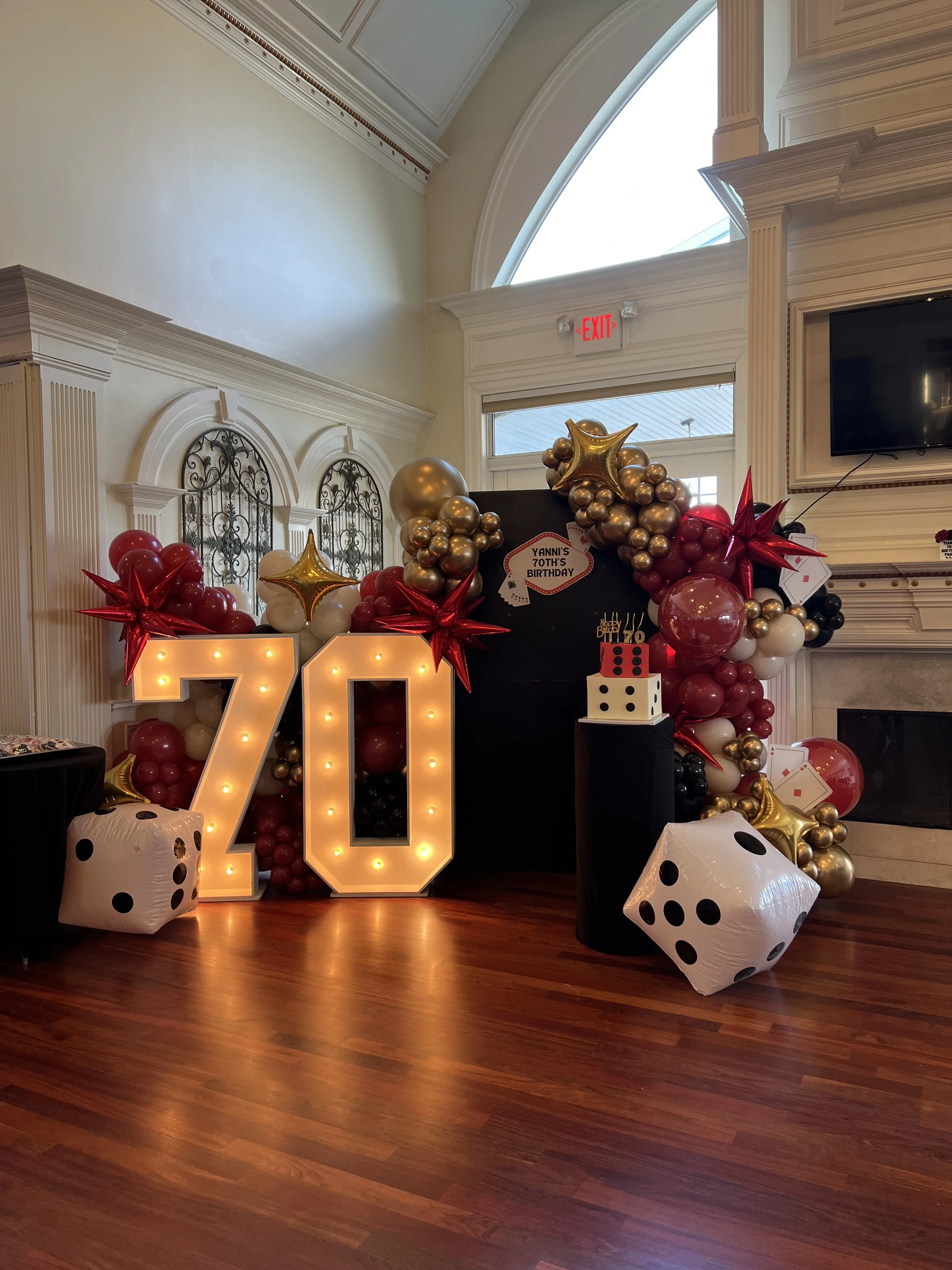 Decorative birthday display with illuminated numbers '70', balloons in red, gold, black, and white, giant dice, and a small birthday cake, celebrating Yanni's 70th birthday.