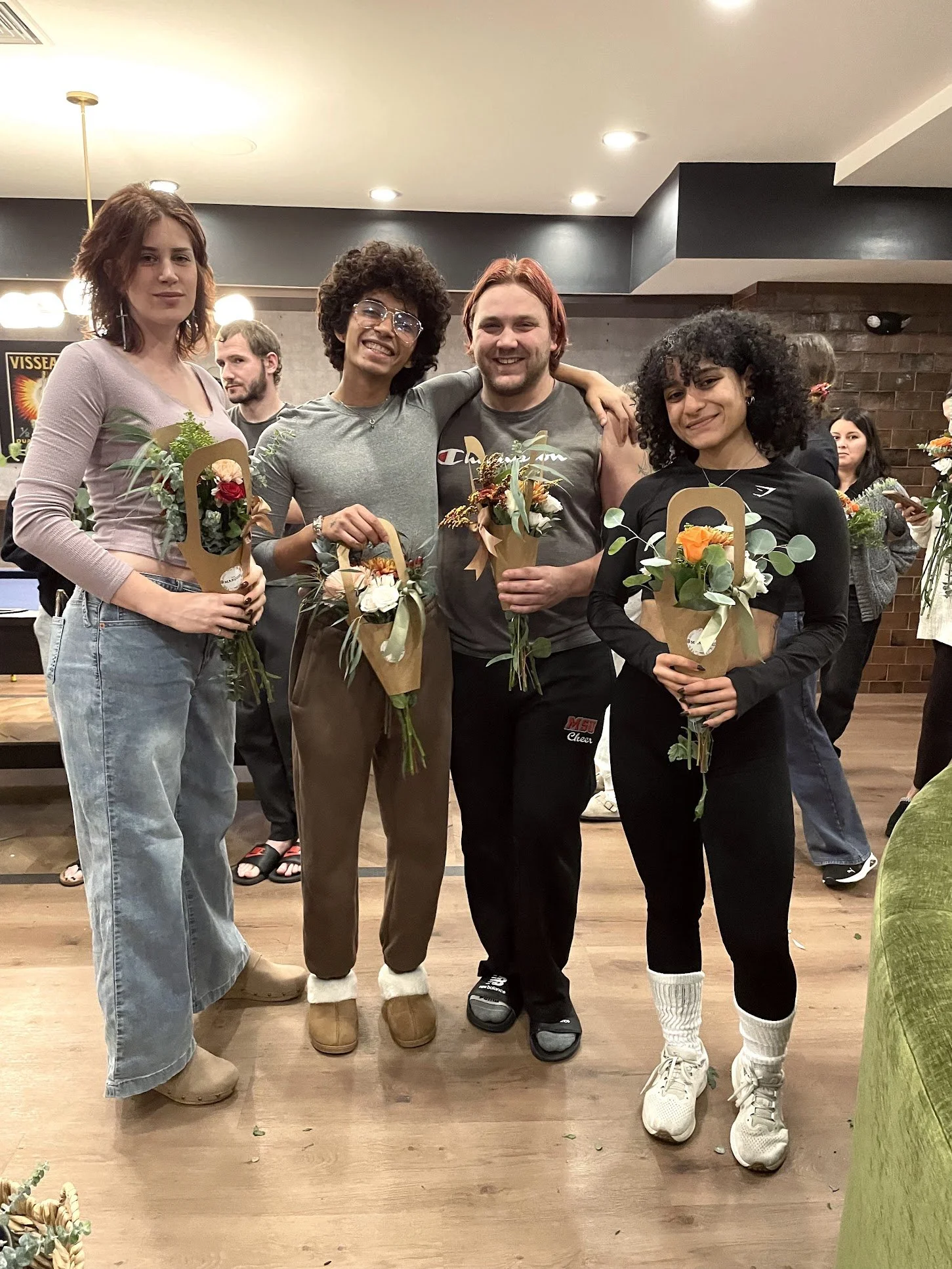 Four young adults standing together indoors, holding flower bouquets, smiling for a photo.