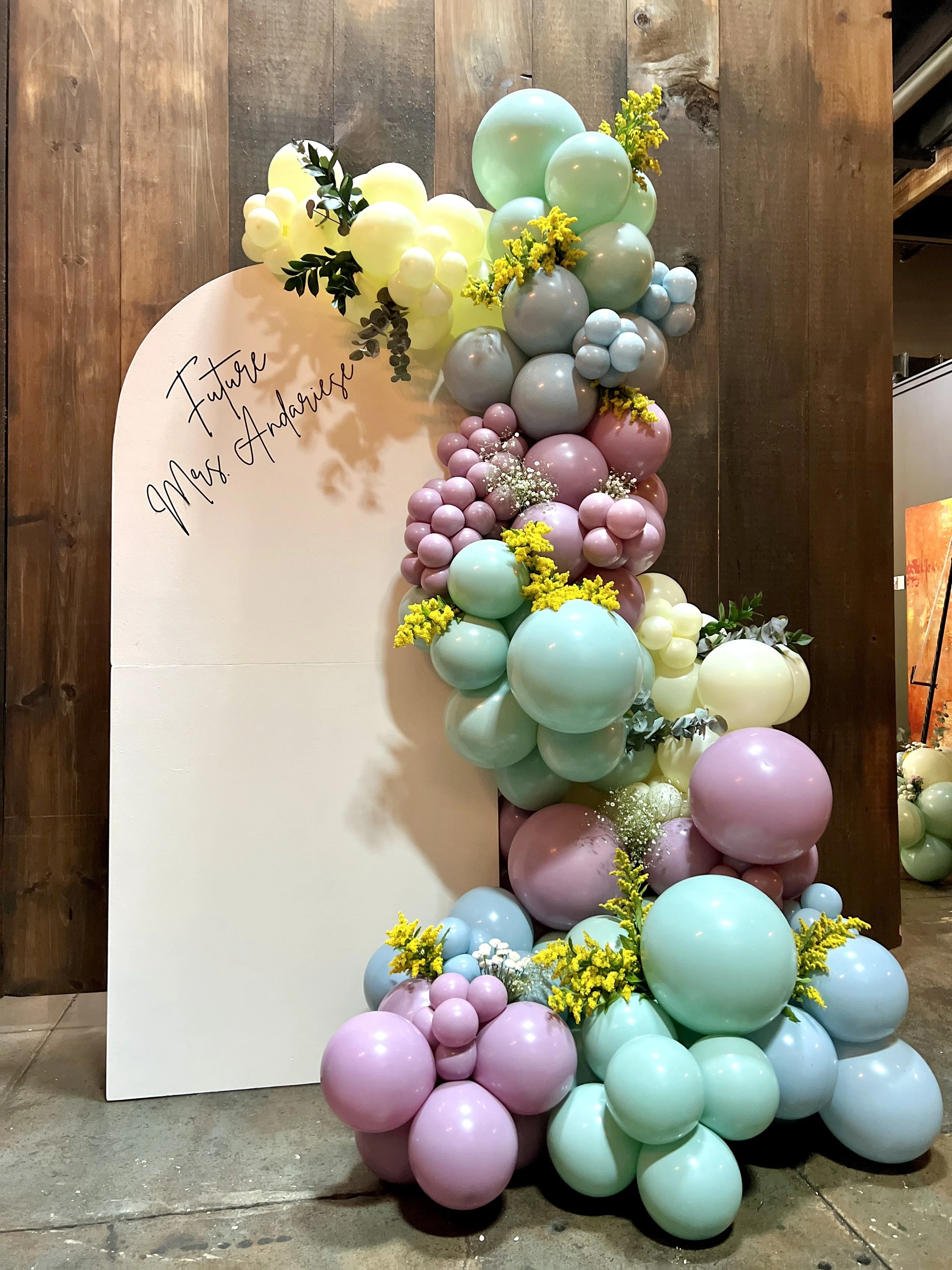 Decorative balloon arrangement with pastel colors including pink, blue, mint, yellow, and white, with flowers and greenery, set beside a white board that has "Future Mrs. Andrade" written on it, on a wooden wall background.