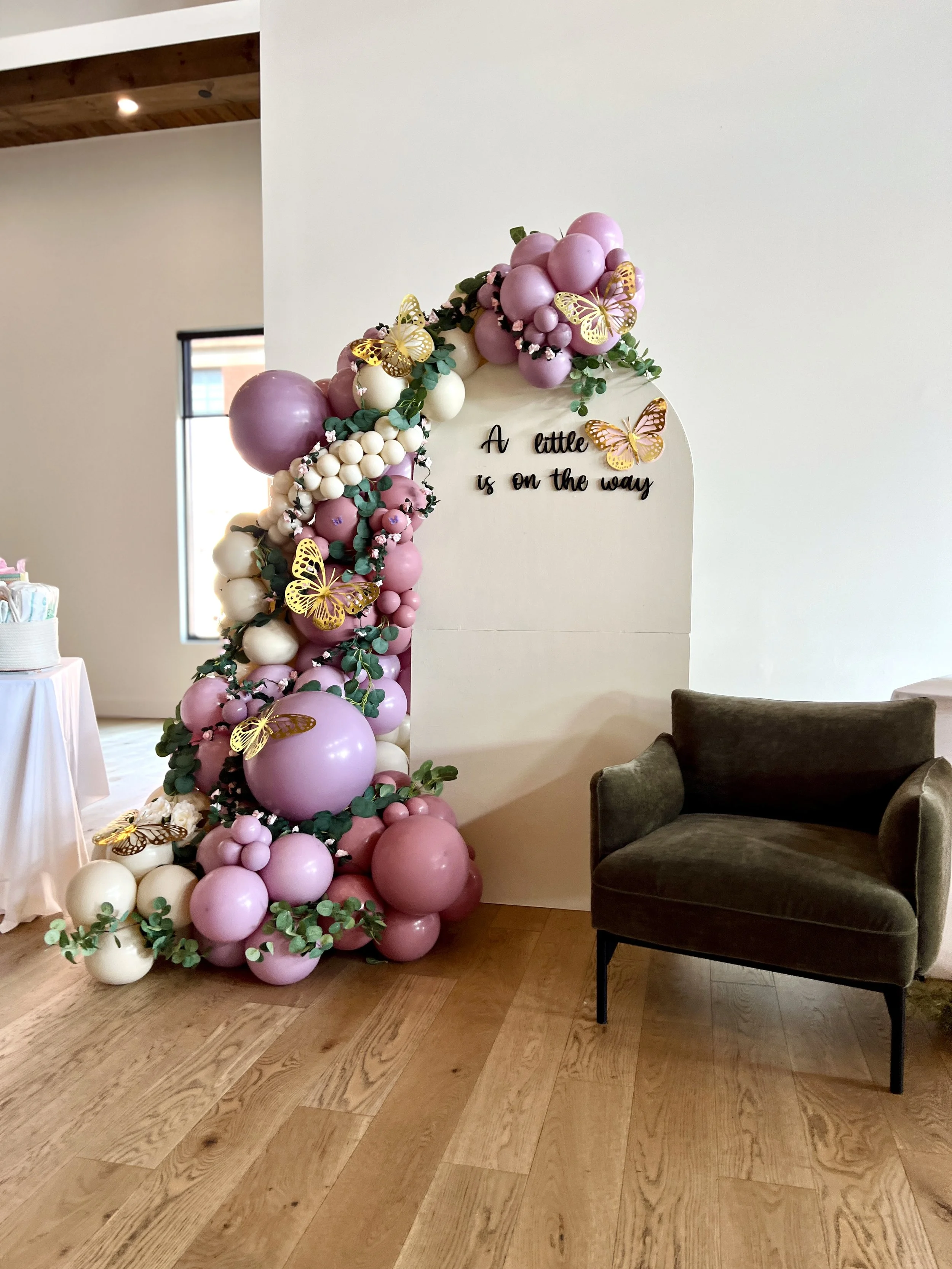 Decorative balloon arrangement with pink, white, and purple balloons, gold butterfly accents, green leaves, and a sign that reads "A little is on the way" in black cursive letters.