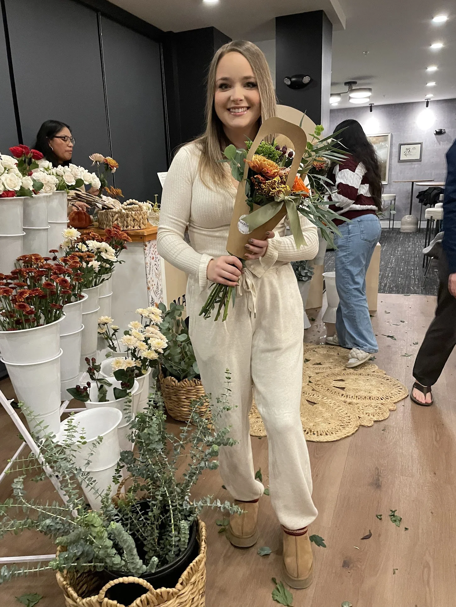 A young woman with long blonde hair smiling and holding a bouquet of flowers, standing inside a flower shop or market with various floral arrangements around her.