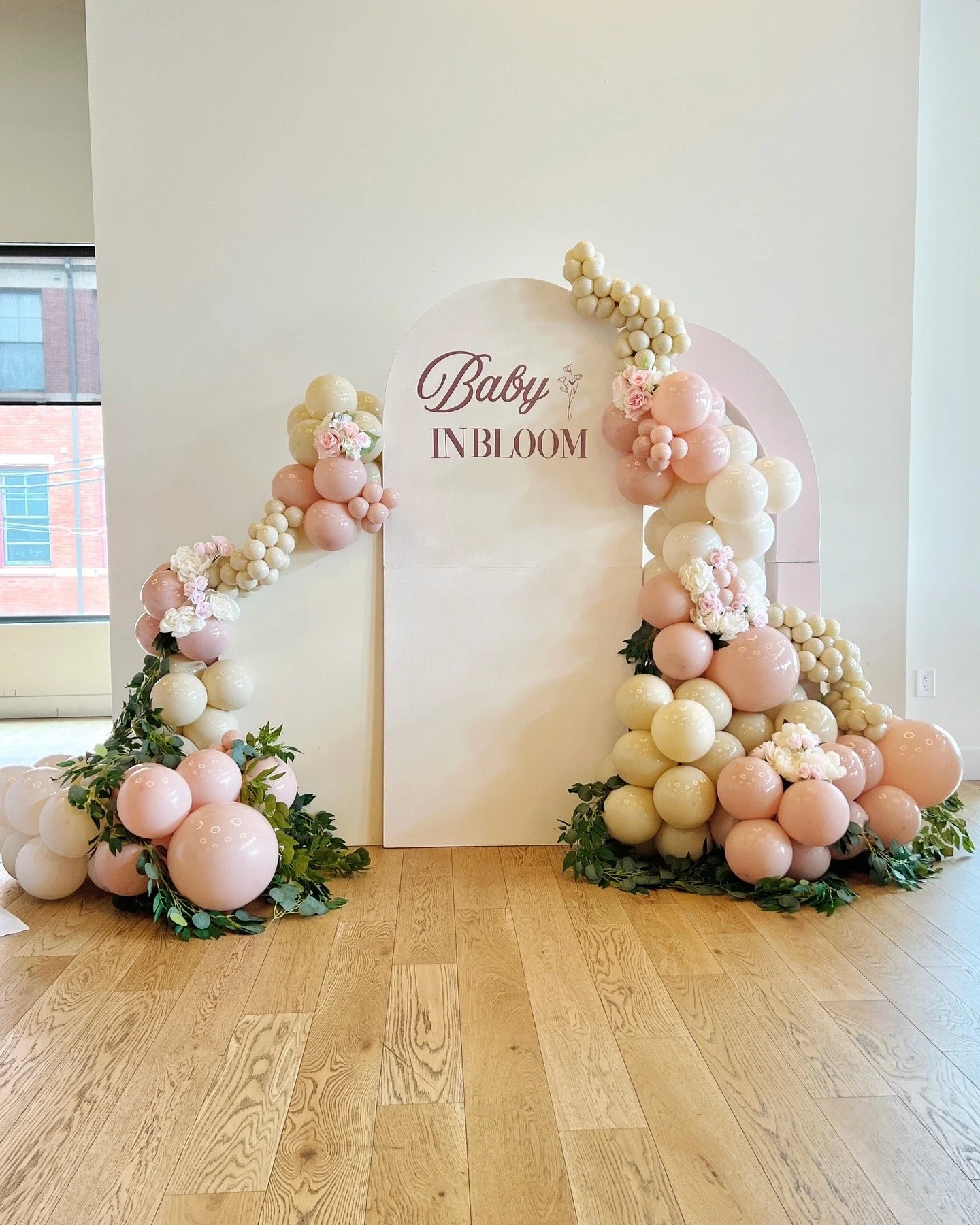 Arched balloon display with pink, white, and cream balloons, decorated with flowers and greenery, with a sign that reads 'Baby InBloom' in elegant script, set against a light-colored wall.