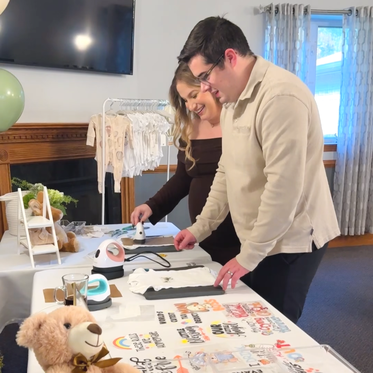 A couple working on heat transfer vinyl crafts at a table, with craft supplies, a stuffed teddy bear, and decorated cloth items displayed.