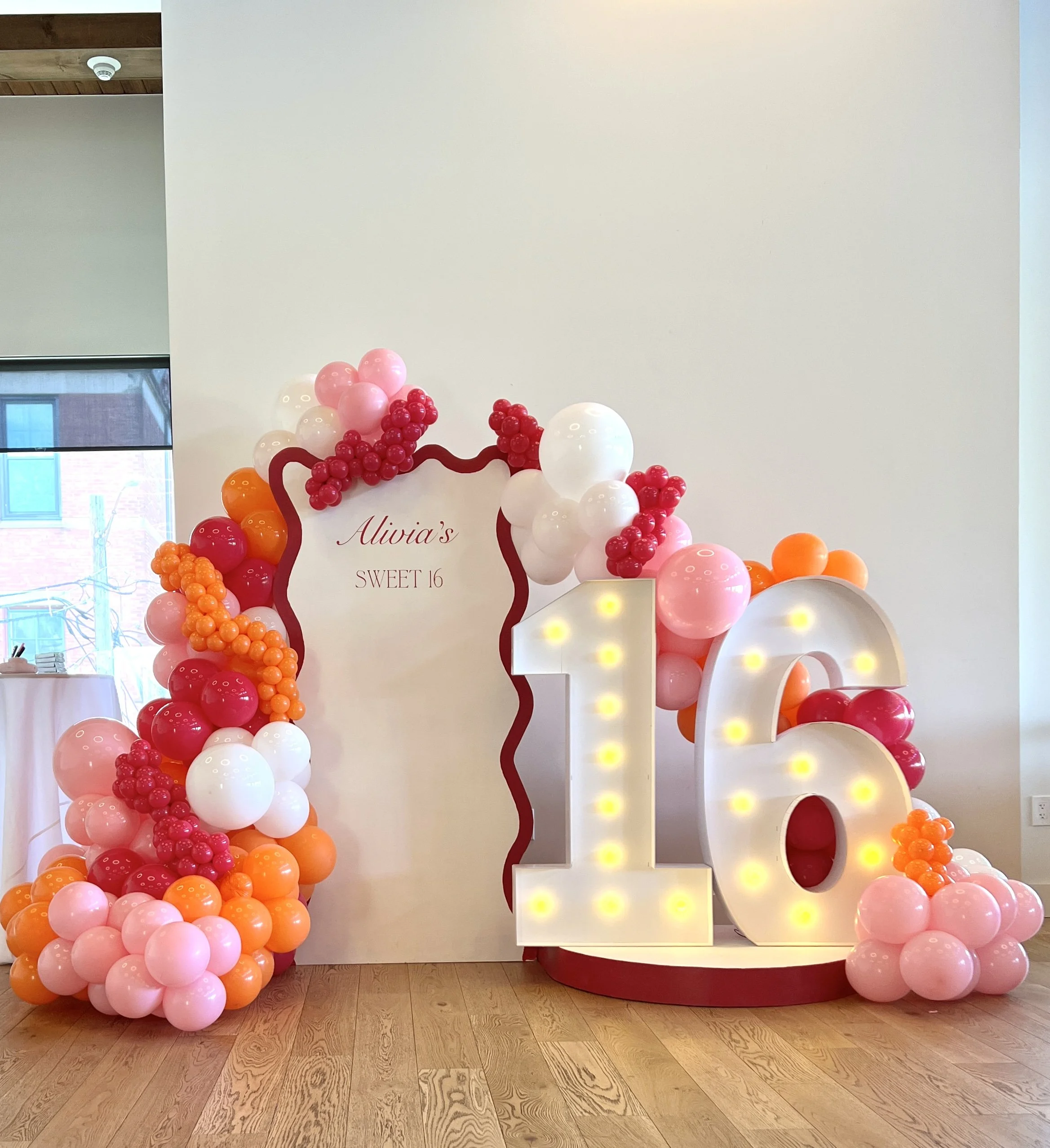 Balloon decoration for a 16th birthday celebration with a large illuminated number 16, pink, white, and orange balloons surrounding a white backdrop that says "Alivia's Sweet 16."